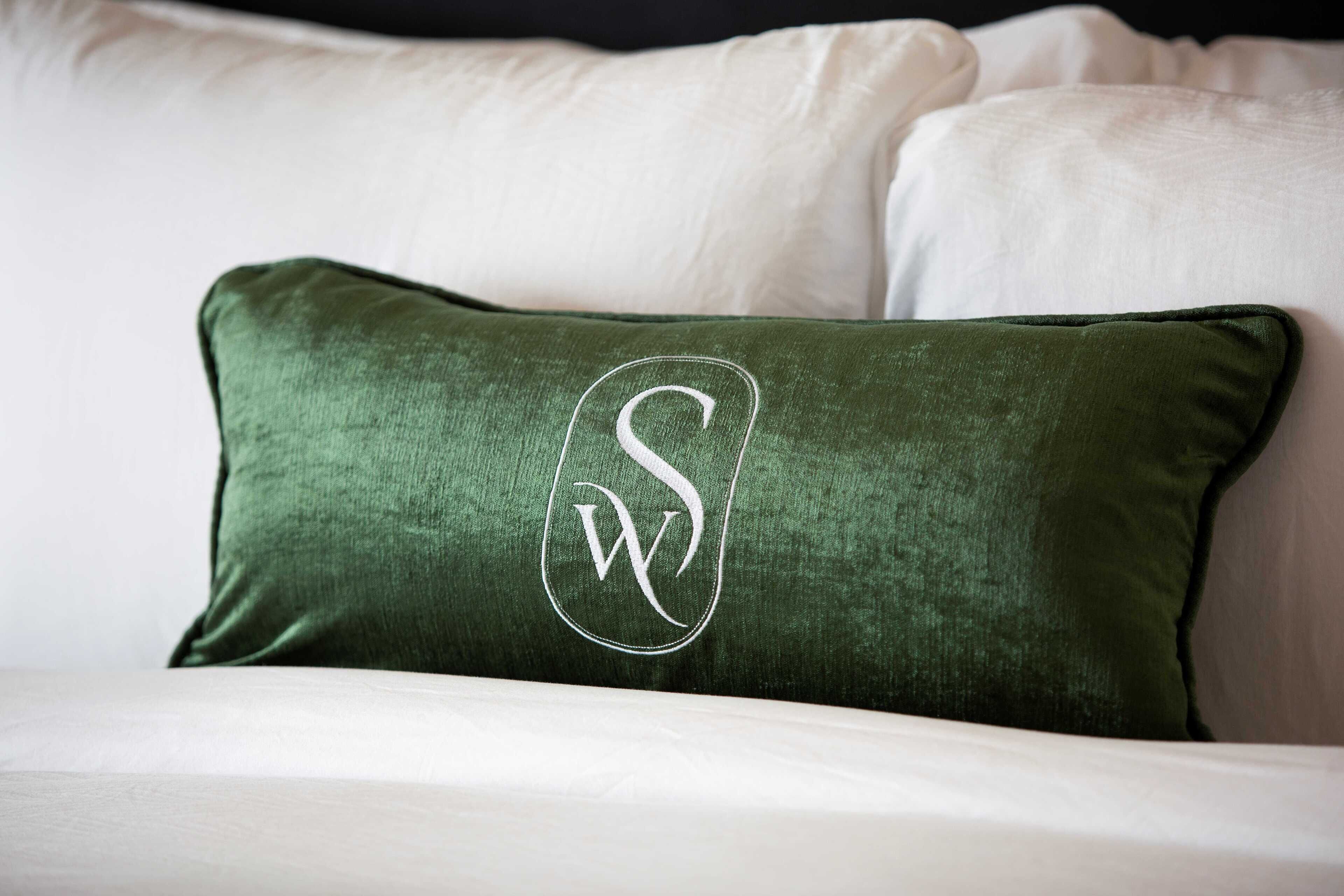 A green pillow with a white embroidered monogram on a white bed.