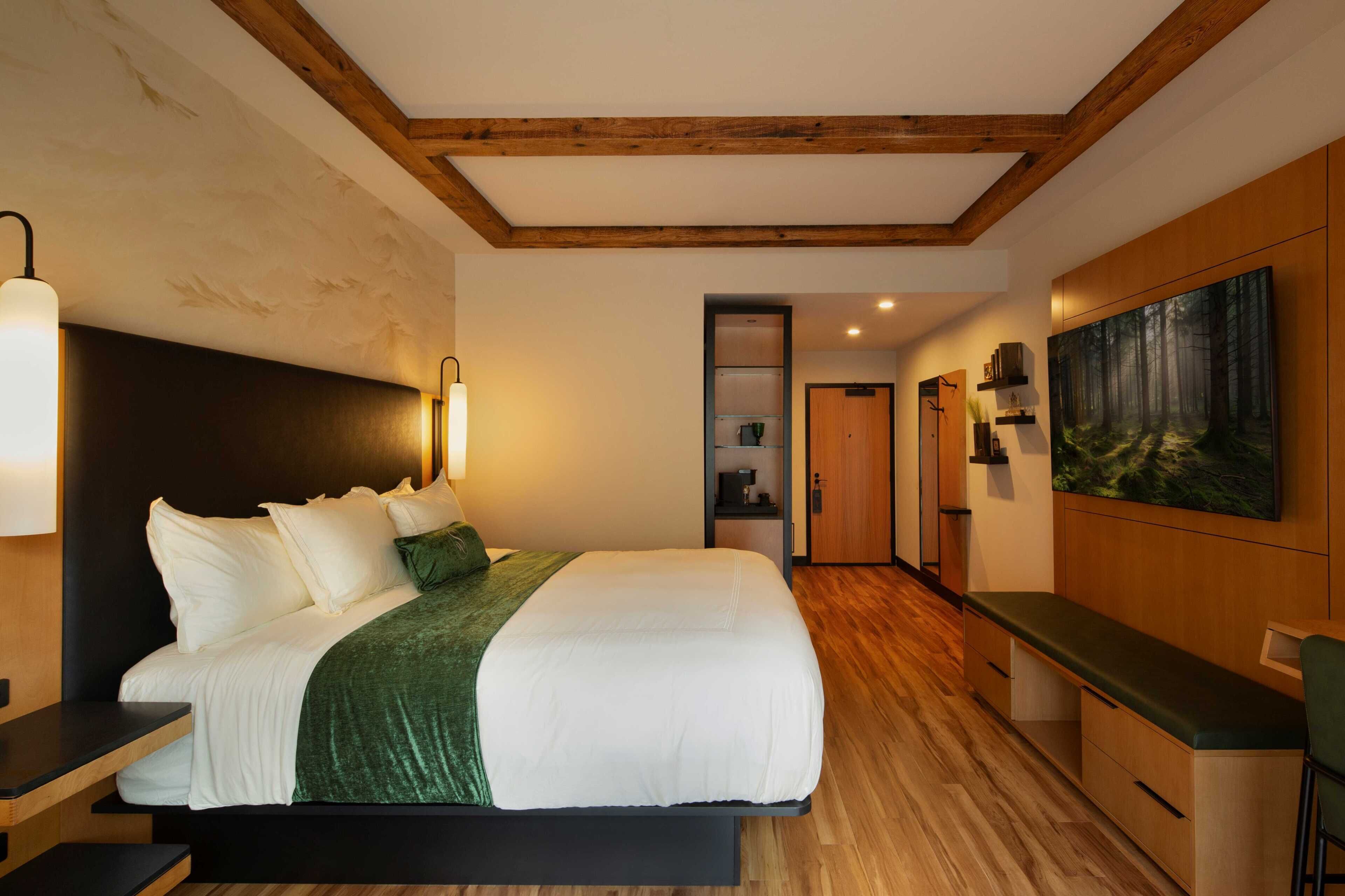 Un dormitorio moderno con una cama grande, vigas de madera en el techo, un plaid verde y un cuadro en la pared.