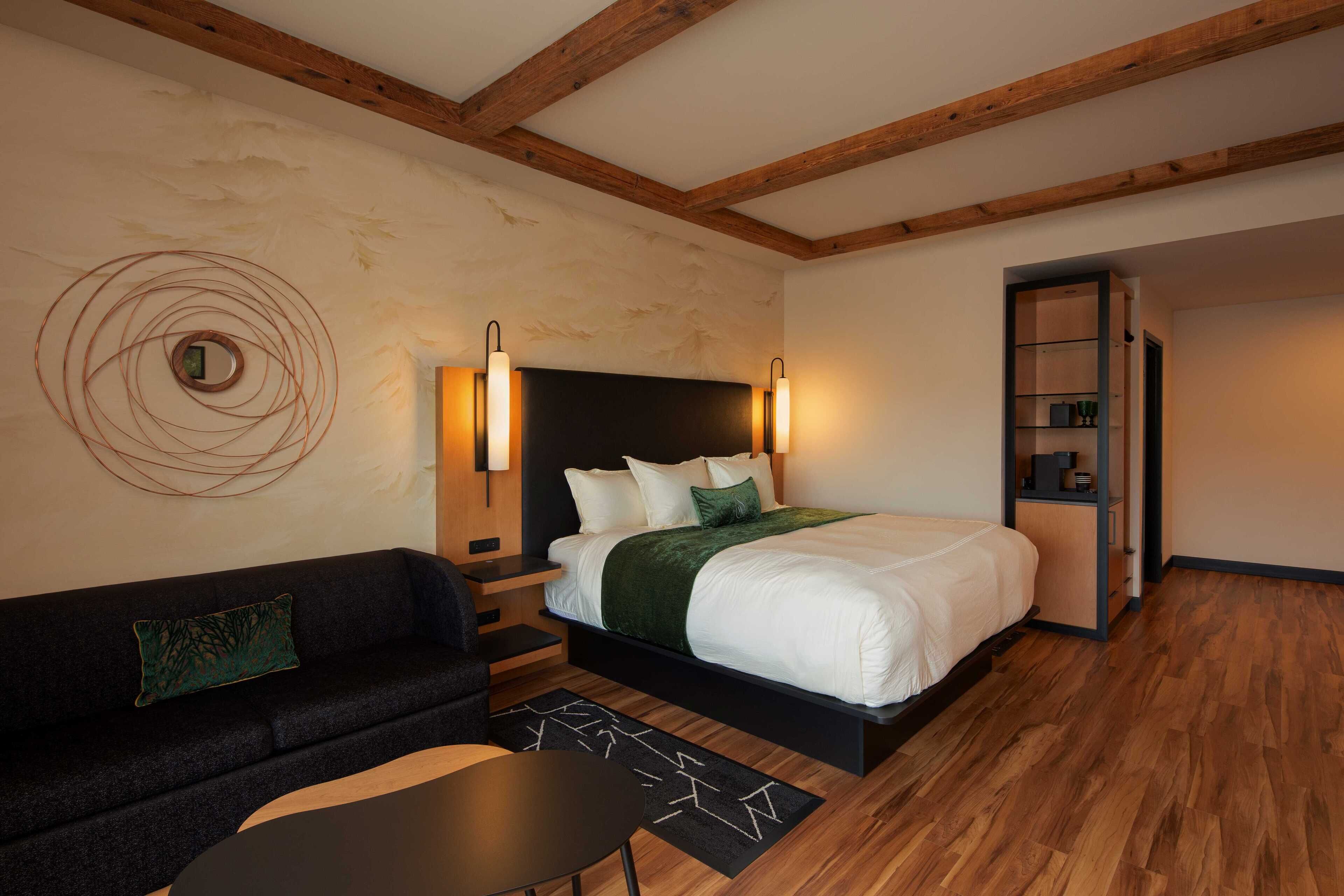 Ein modernes Hotelzimmer mit Holzboden, einem großen Bett mit weißer Bettwäsche, einer schwarzen Couch, einem kleinen Tisch und einer Wandleuchte.