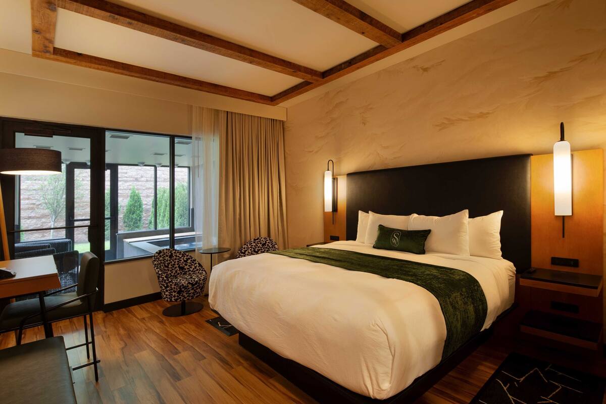 A hotel room with a large bed, a desk, and a chair. There is a window with curtains, a lamp, and a wall-mounted light fixture.