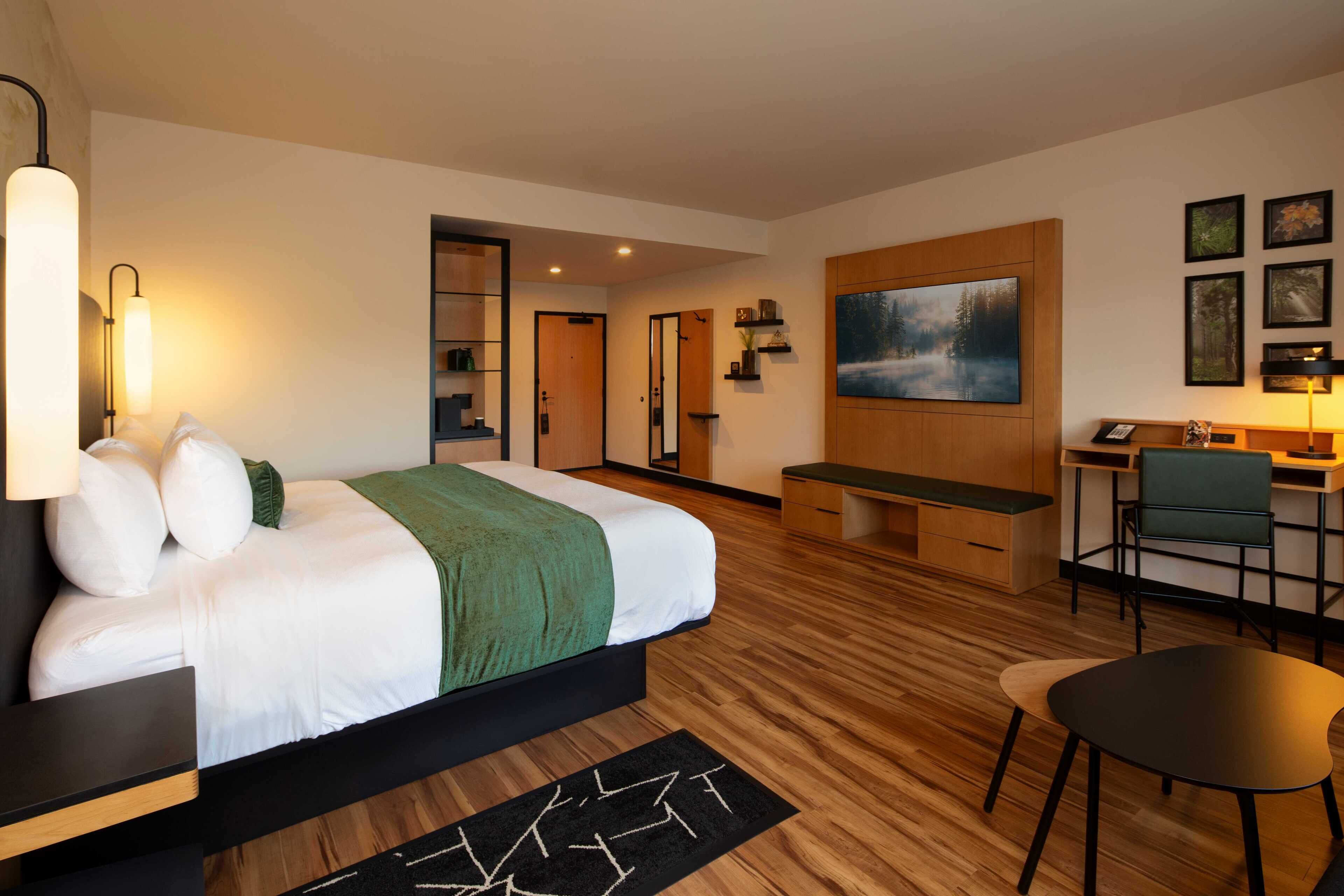 A modern hotel room with a large bed, a desk, and a chair. There is a painting on the wall and a desk lamp.