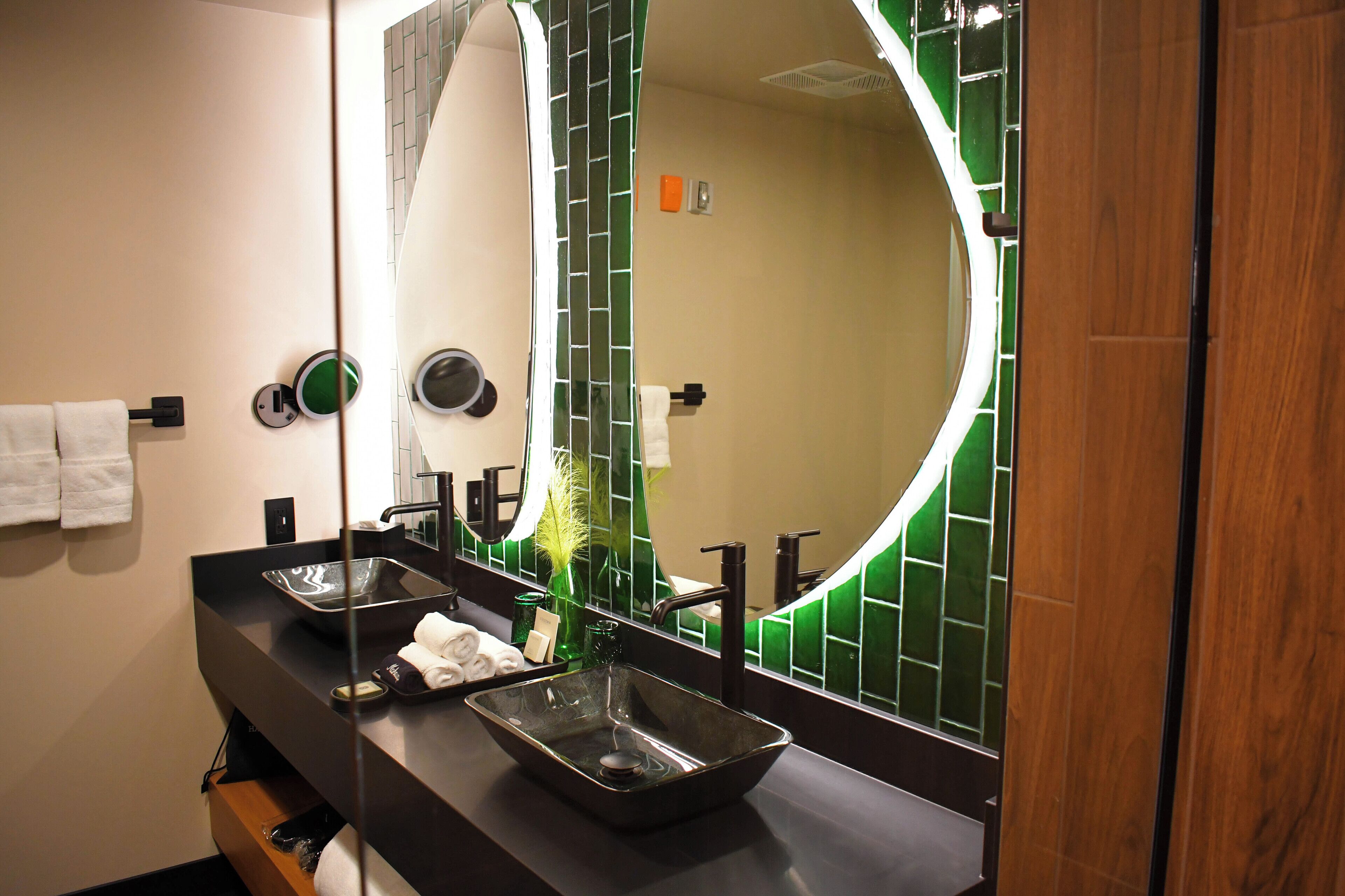A modern bathroom with a black countertop, a large mirror, and a green-tiled wall.