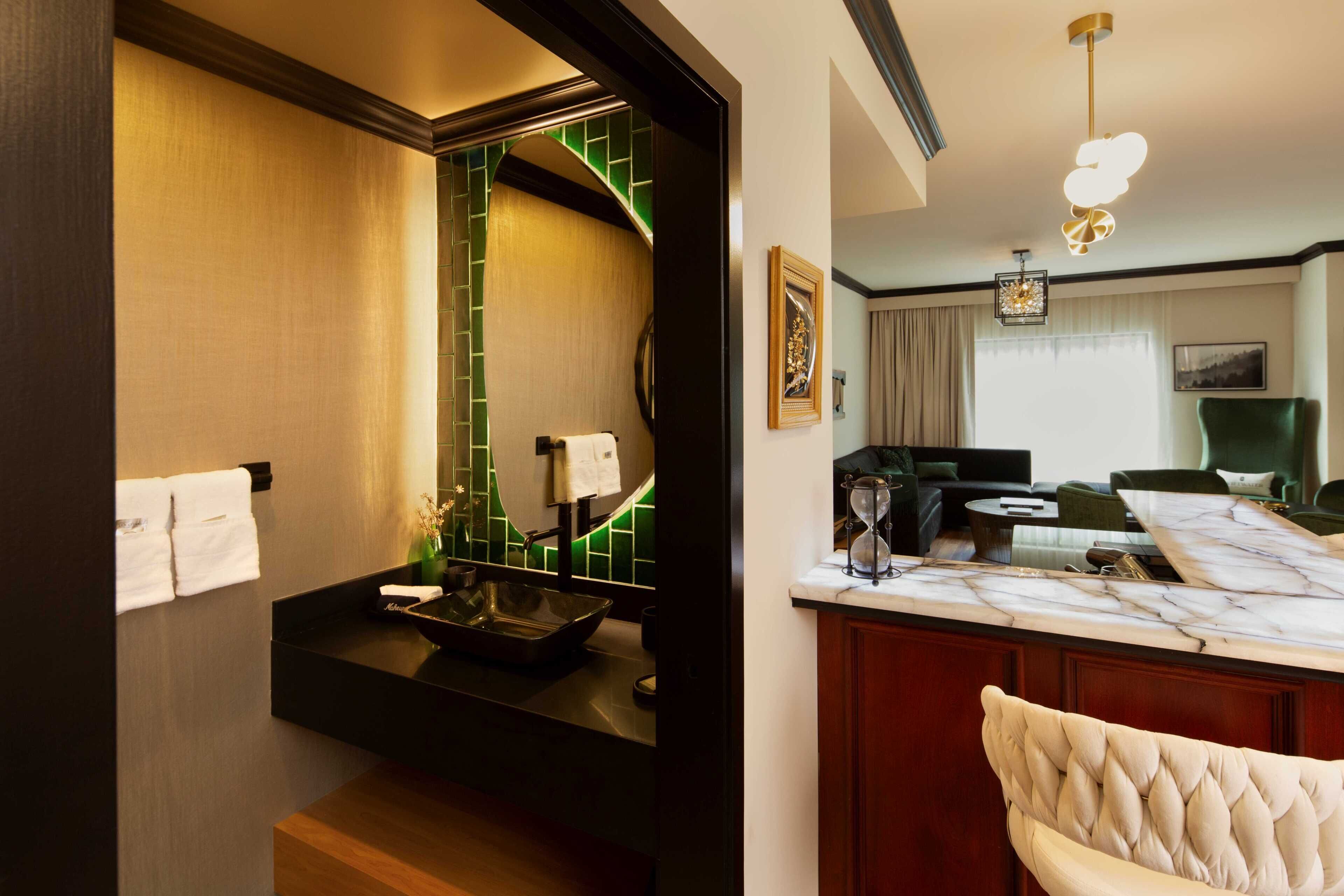 Una habitación de hotel con un baño moderno, una zona de comedor con una mesa de mármol y una sala de estar con un sillón verde y un sofá.
