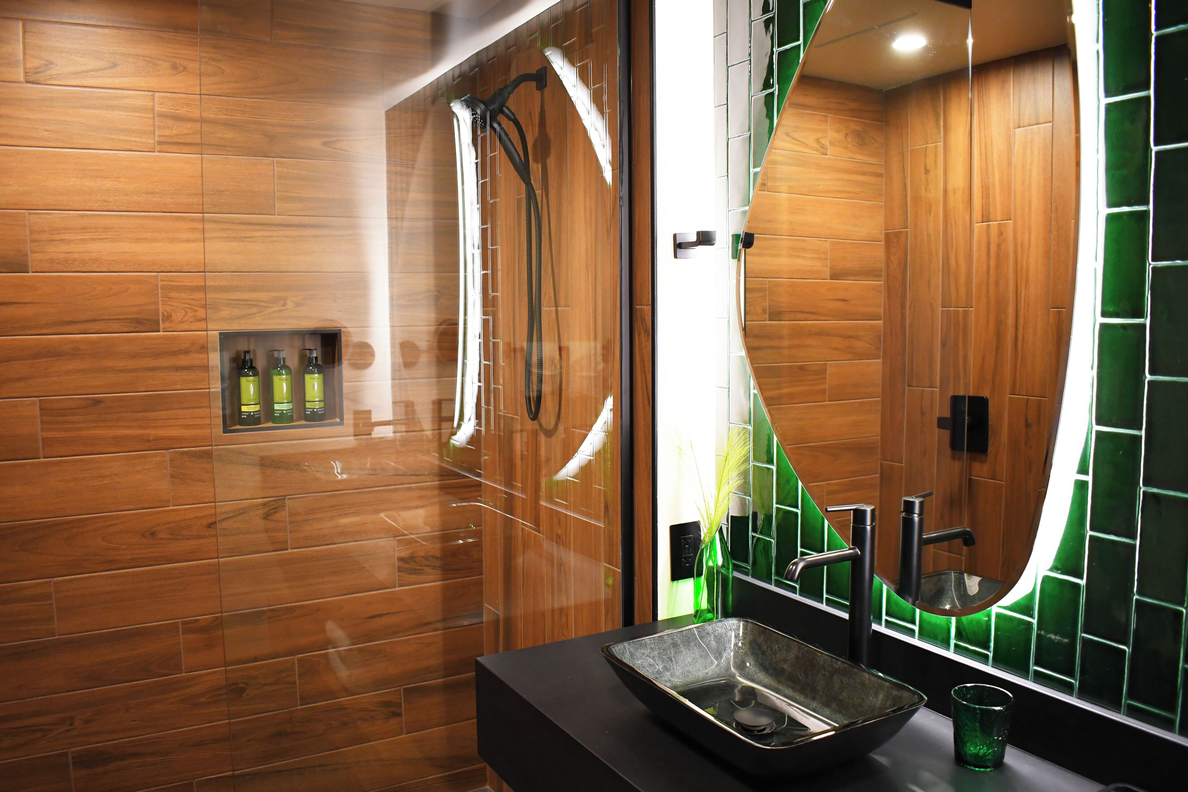 A modern bathroom with wooden walls, a black sink, and a green-tiled mirror.
