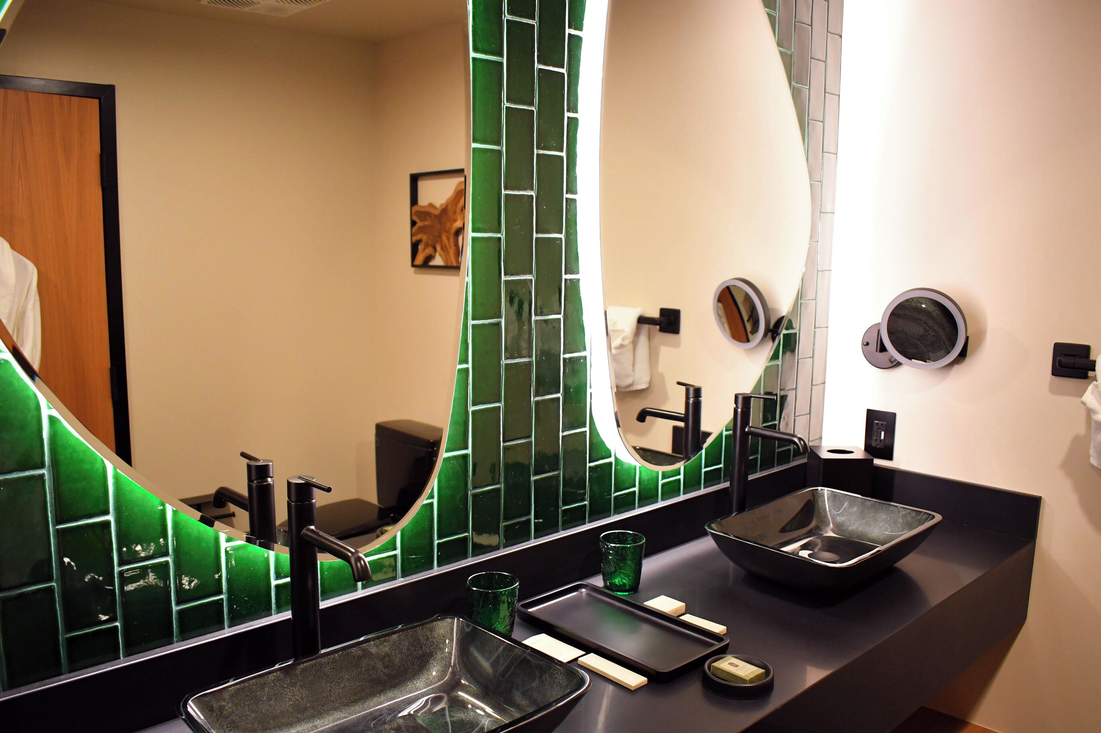 A bathroom with a black countertop, two sinks, a green-tiled wall, and a mirror.