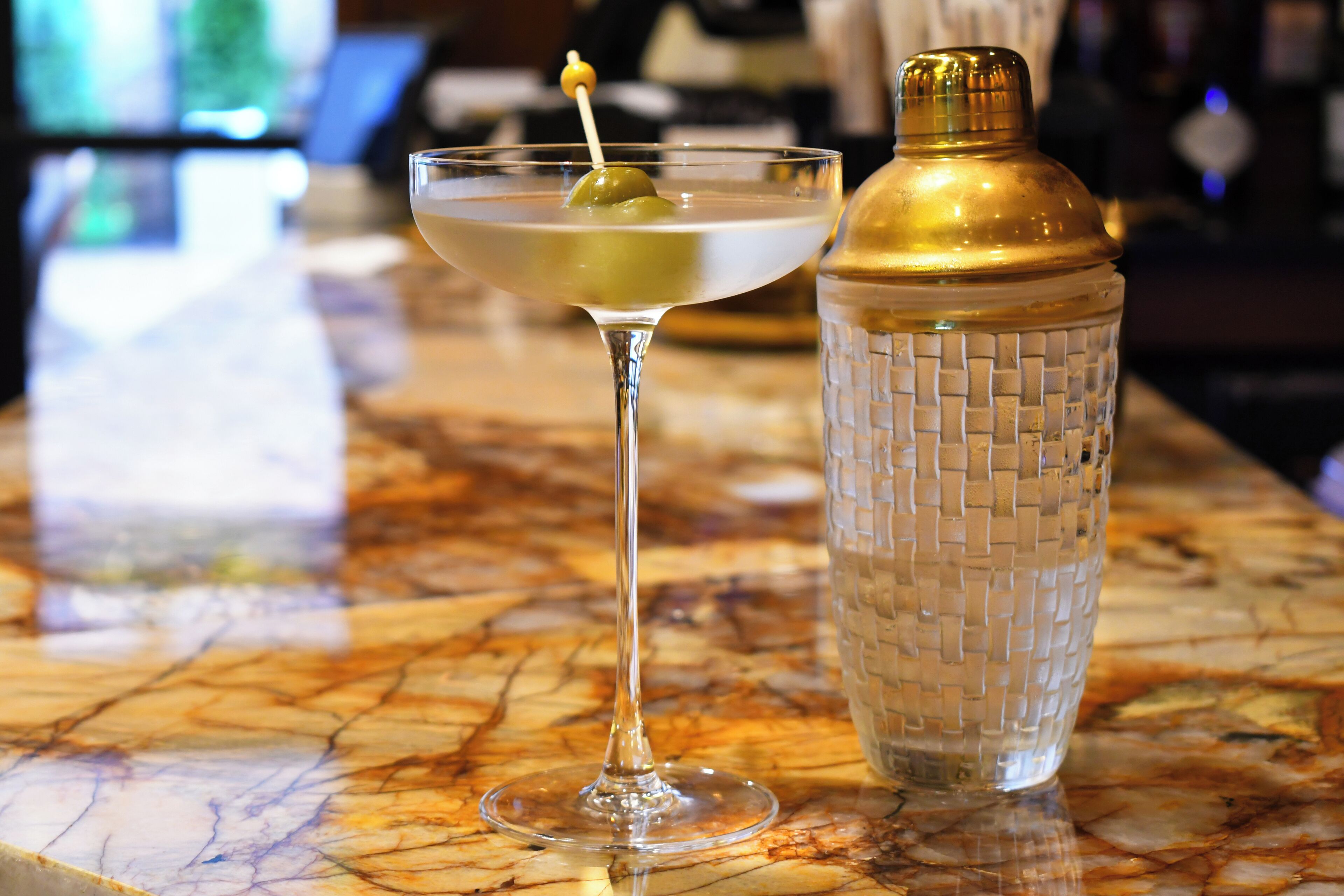 Una copa de martini con aceituna sobre una barra de mármol junto a un coctelero texturizado.
