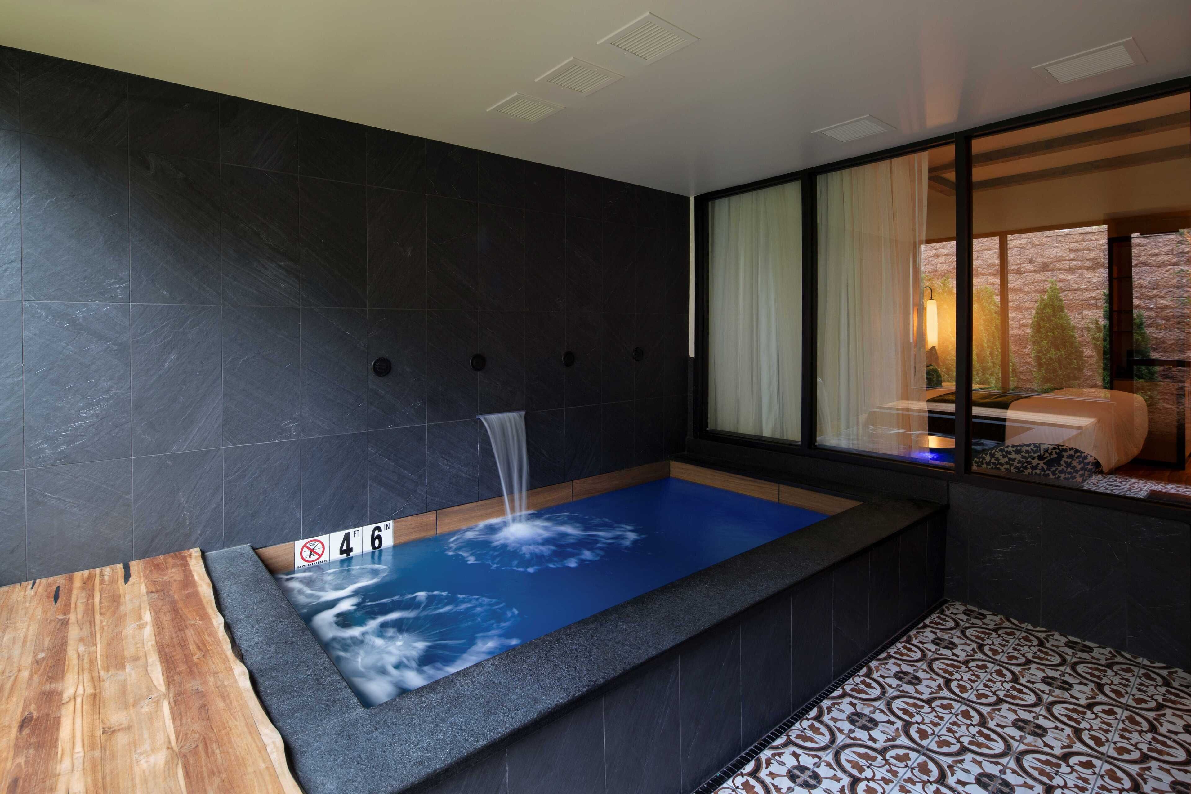 A modern bathroom with a blue-tiled bathtub, a wooden bench, and a large window with curtains.