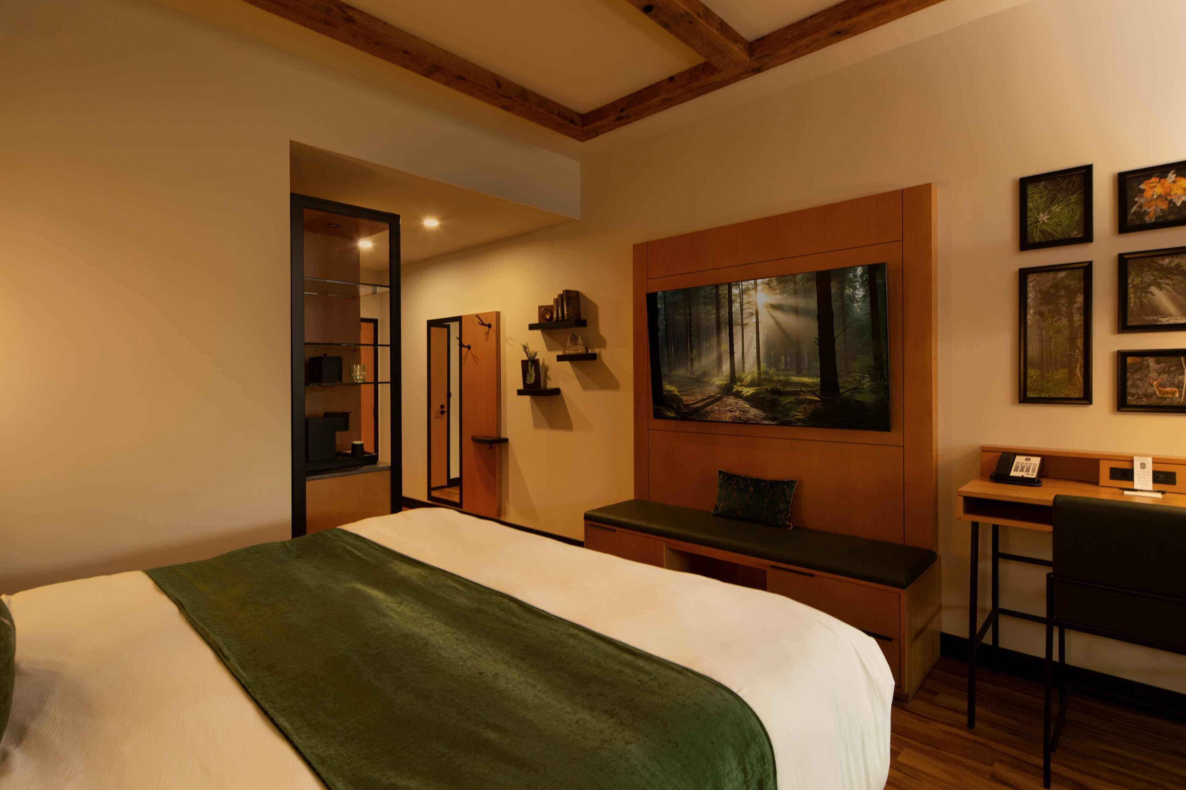Quarto de hotel com uma cama grande, uma escrivaninha, uma cadeira, um quadro de uma floresta e um espelho.