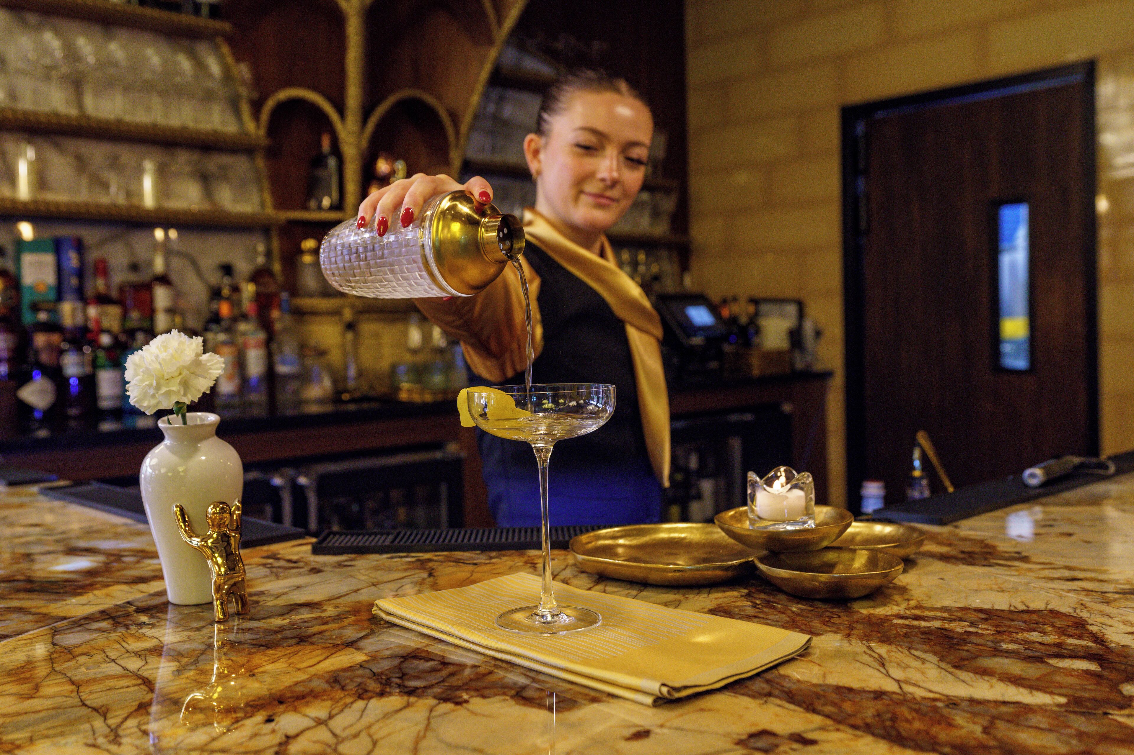 Una zona de bar con encimera de mármol, un jarrón blanco con una flor, una figurita dorada y una copa de martini.