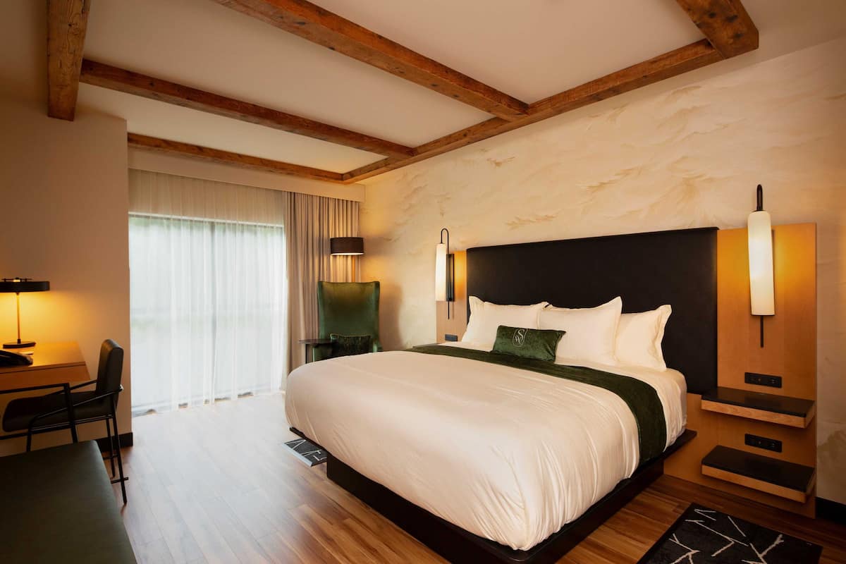 A large bed with a dark headboard, a bedside table, a lamp, and a chair in a room with wooden beams and a large window.