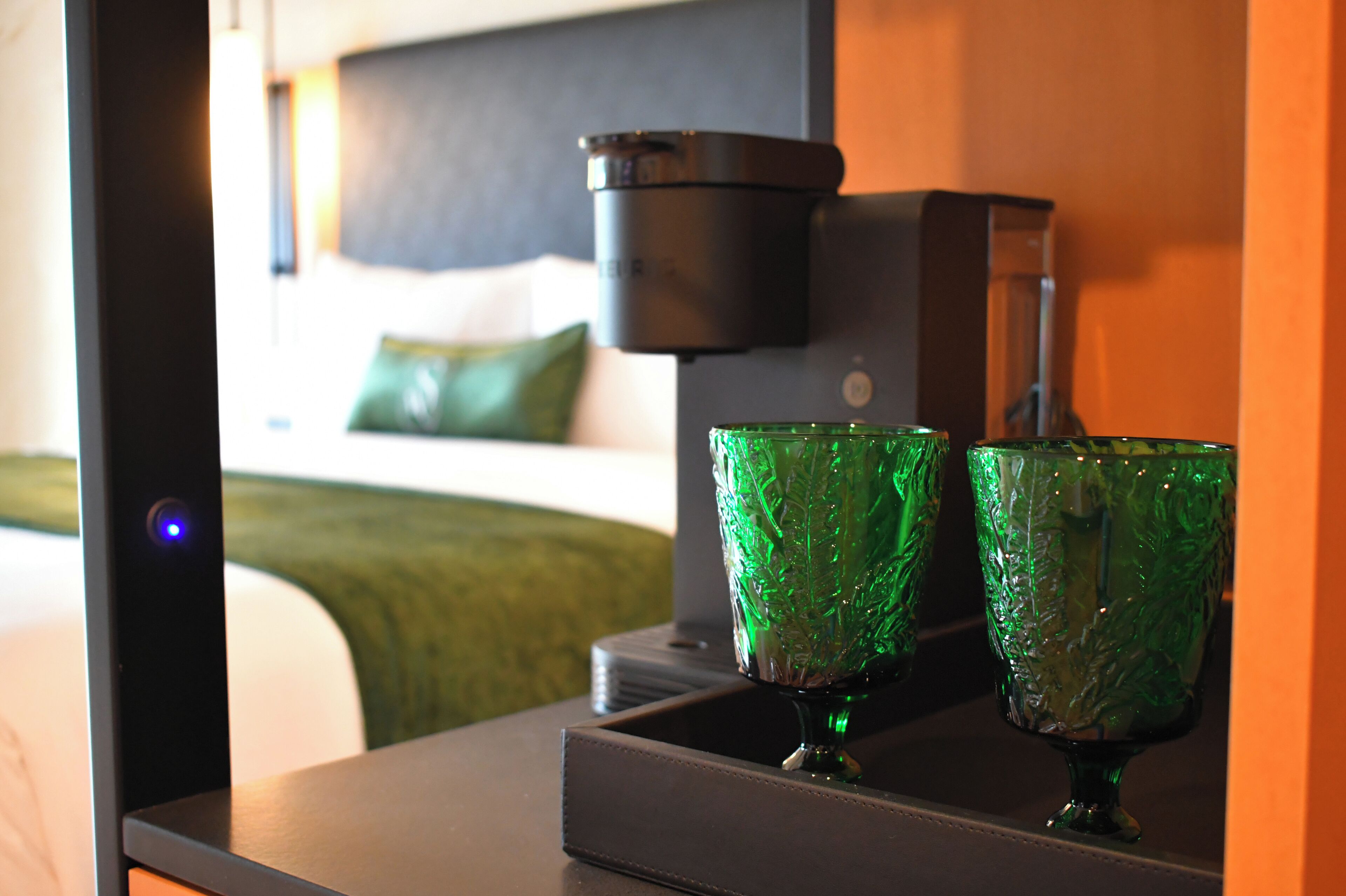 A hotel room with a bed, a bedside table with two green glasses, a black lamp, and a wall-mounted device.