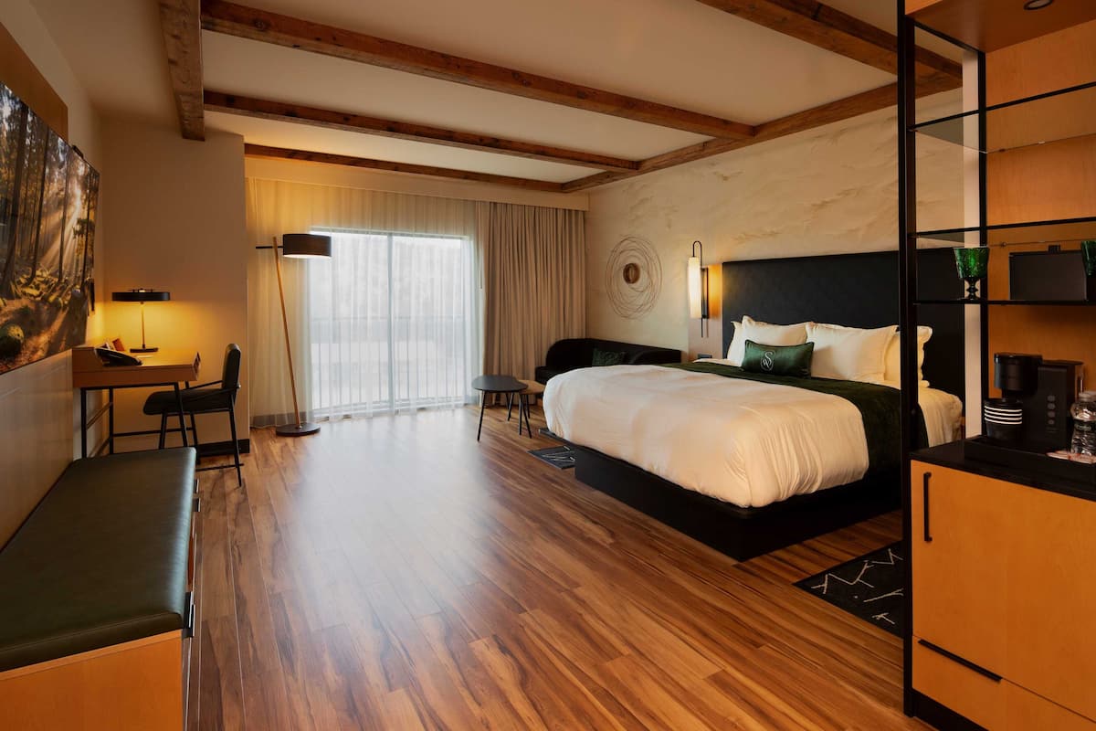 A modern hotel room with a large bed, a desk, and a chair. There is a large window with curtains, a painting on the wall, and a wooden floor.
