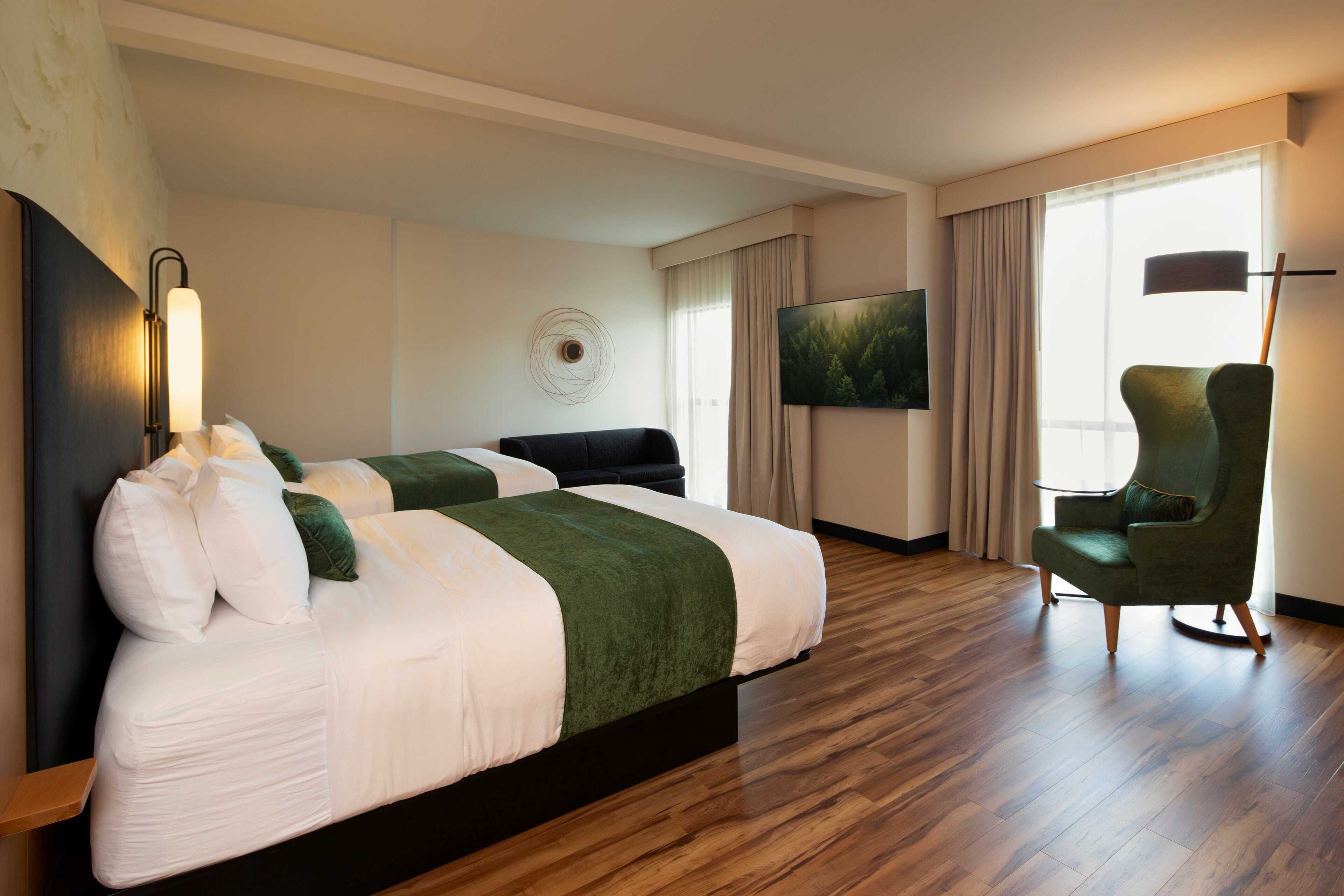 Una habitación de hotel con dos camas, un sillón verde, un televisor de pantalla plana y un ventanal con cortinas.