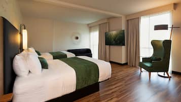 Una habitación de hotel con dos camas, un sillón verde, un televisor de pantalla plana y un ventanal con cortinas.