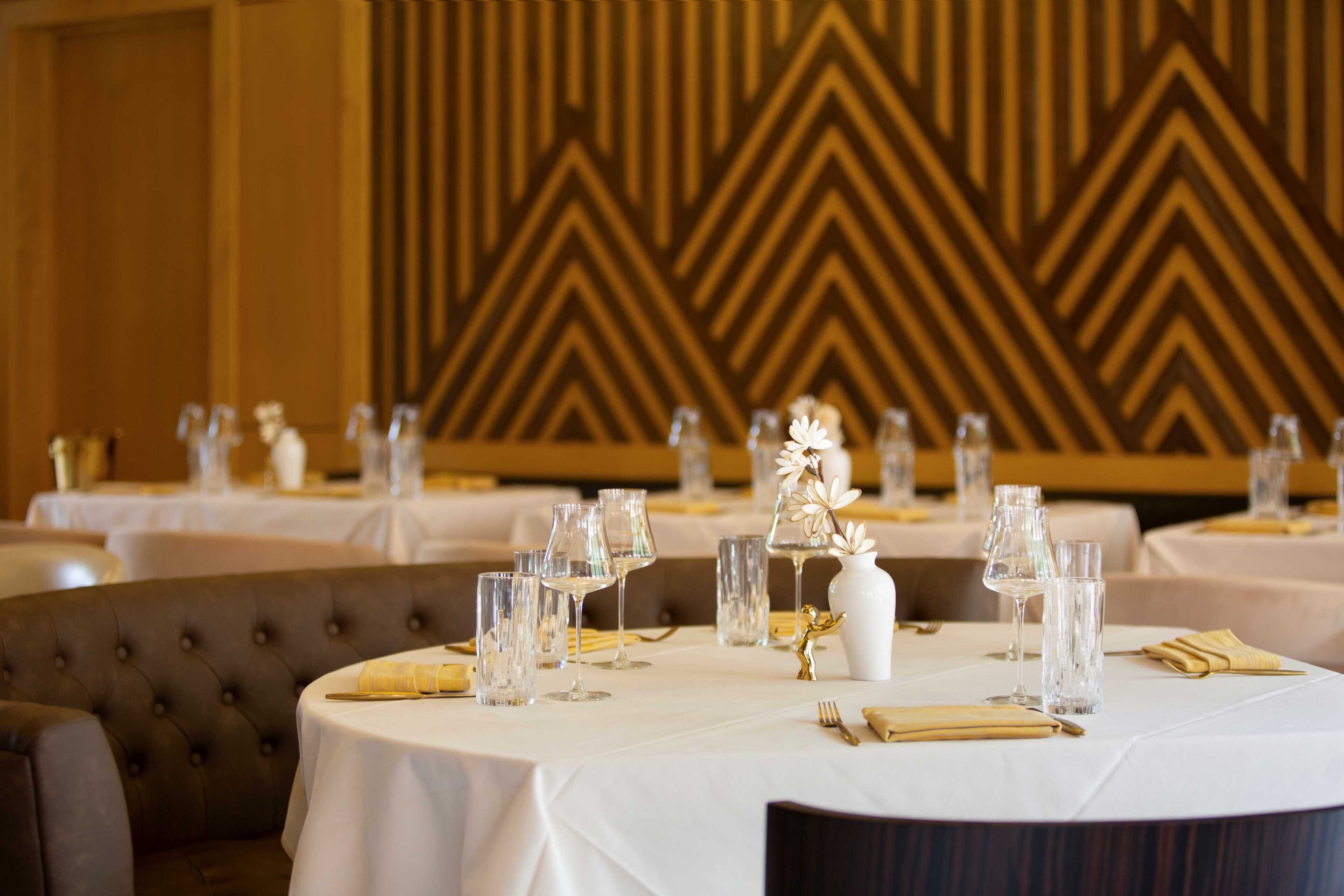 A dining area with round tables set for a meal, featuring white tablecloths, clear glassware, and gold napkins.