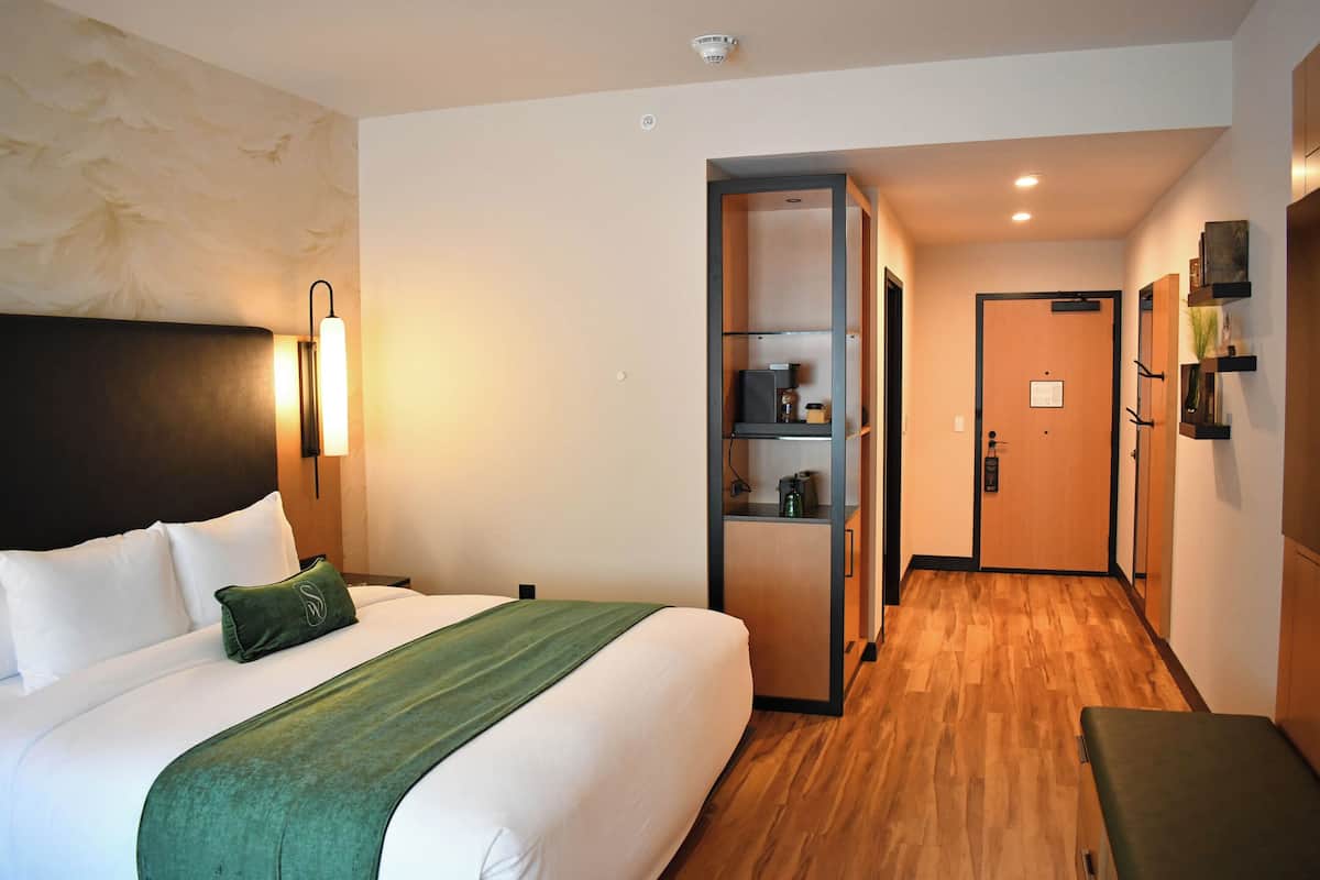 A hotel room with a bed, bedside table, wall-mounted lamp, and a wooden floor.