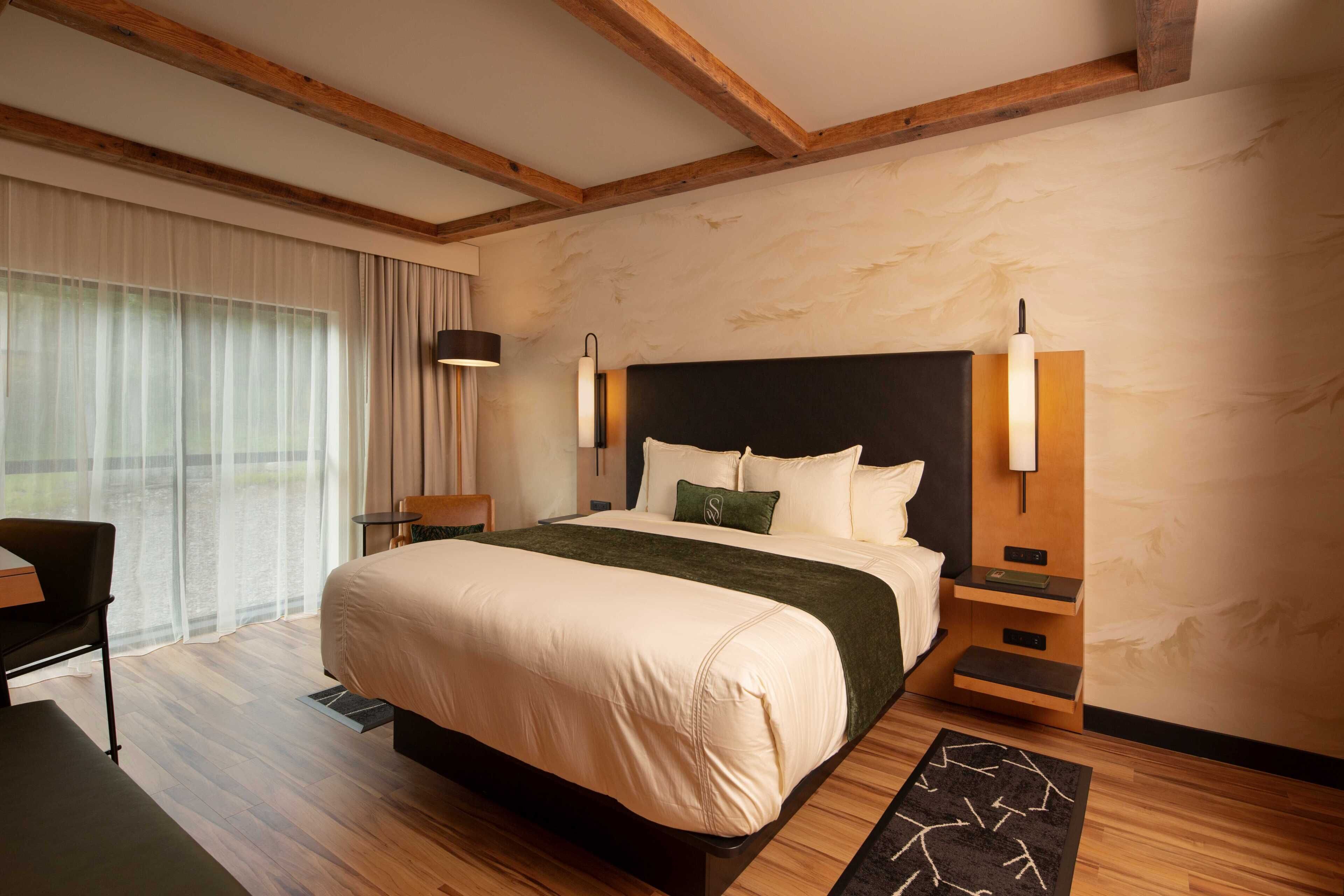 A modern hotel room with a large bed, wooden beams on the ceiling, a desk, and a chair.