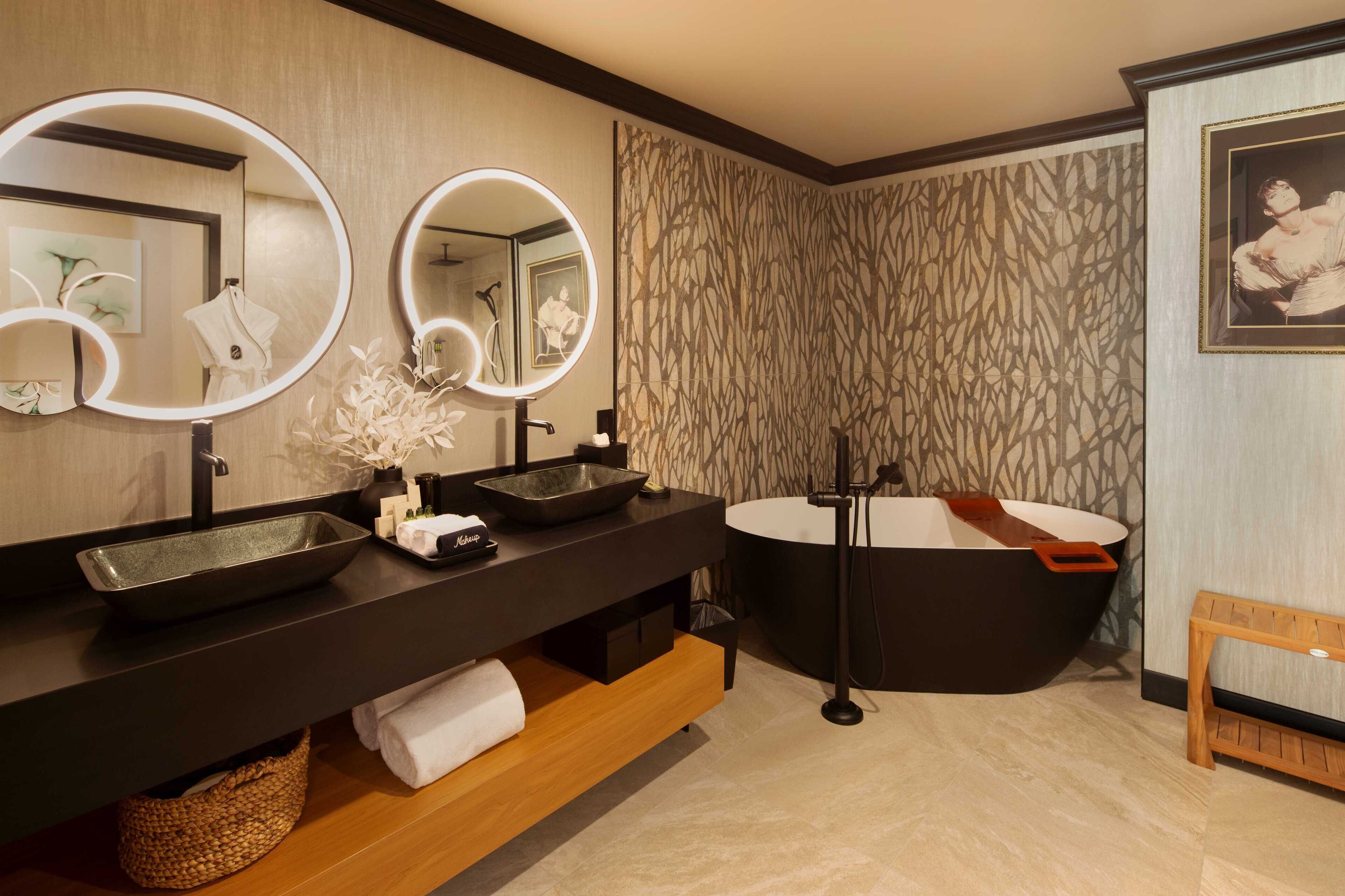 A modern bathroom with a black vanity, two round mirrors, a black bathtub, and a wooden bench.