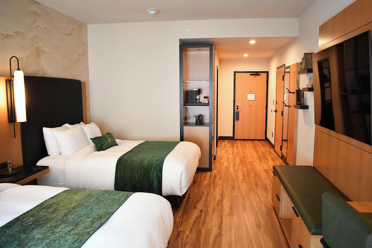 A hotel room with two beds, a desk, a bench, a wardrobe, and a bathroom visible through an open door.