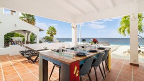Un área de comedor al aire libre techada con terraza y vistas al mar, con una mesa de madera dispuesta para comer y panorama oceánico.