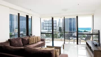Una sala de estar moderna con un sofá grande, una mesa de centro y un mueble para televisión. La habitación tiene ventanas amplias con vistas a rascacielos y un balcón.