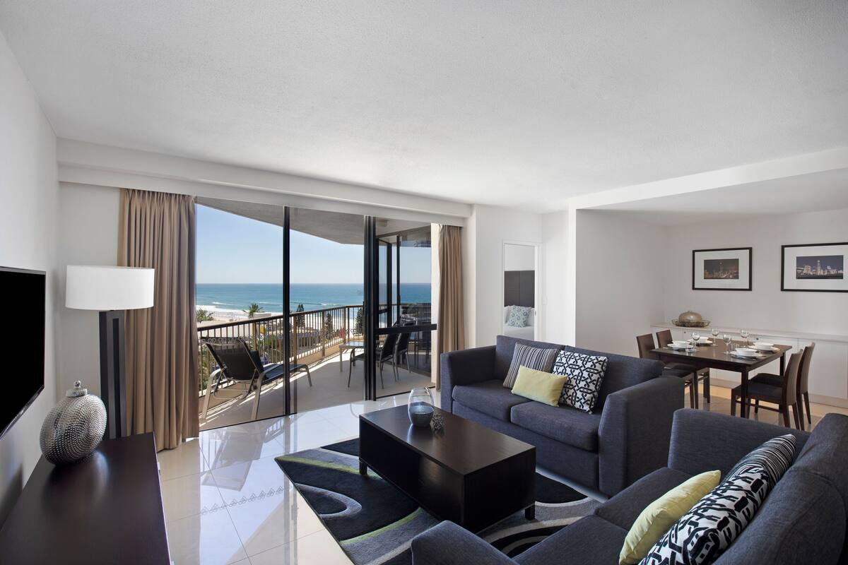 A modern living room with a sofa, coffee table, and a dining area. A large window offers a view of the ocean.