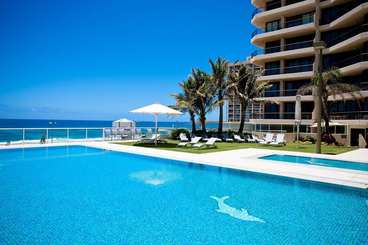 Una piscina azul cristalina con sillas de descanso y sombrillas, un edificio con balcones y vistas al océano.