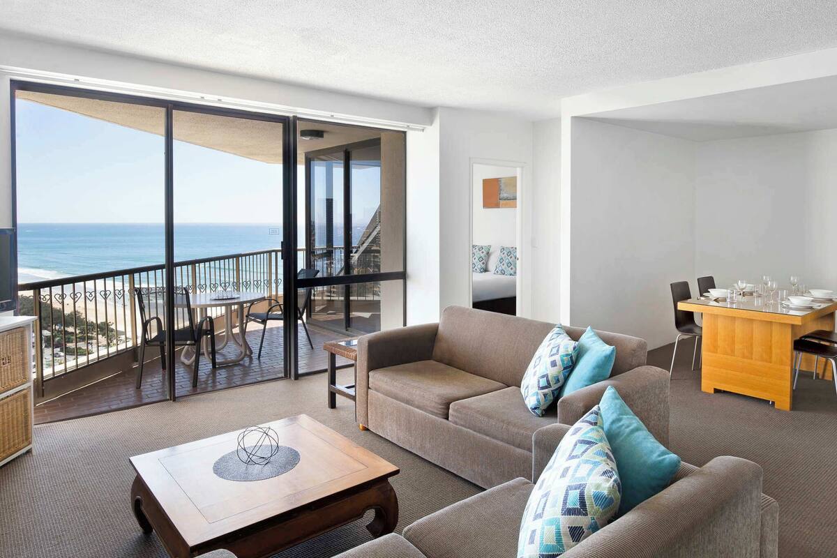 A spacious living area with a balcony overlooking the ocean, a dining table set, and a sofa with patterned cushions.