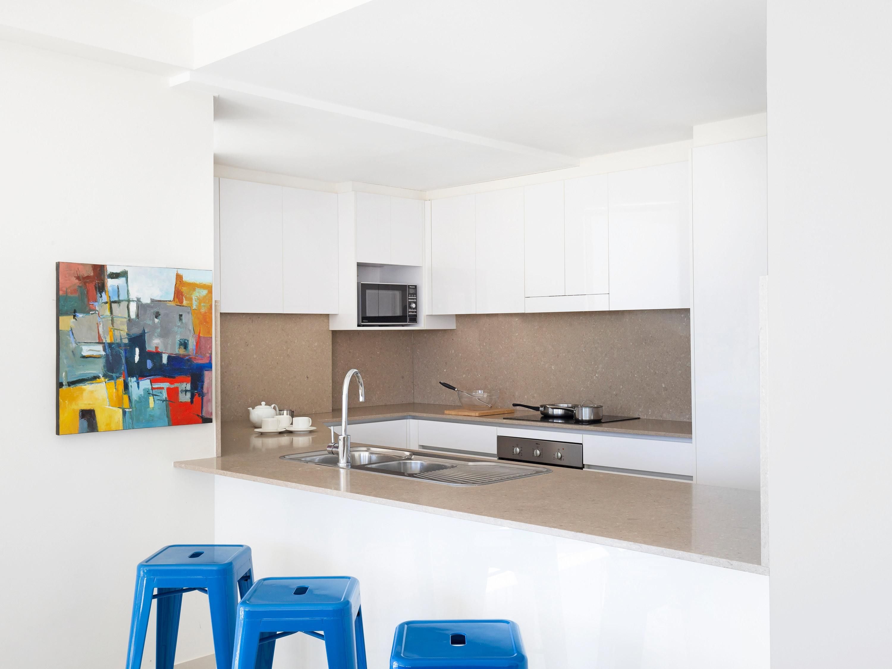 Una cocina moderna con armarios blancos, un lavamanos de acero inoxidable, un microondas y una estufa. Hay dos taburetes azules y un cuadro abstracto de colores en la pared.