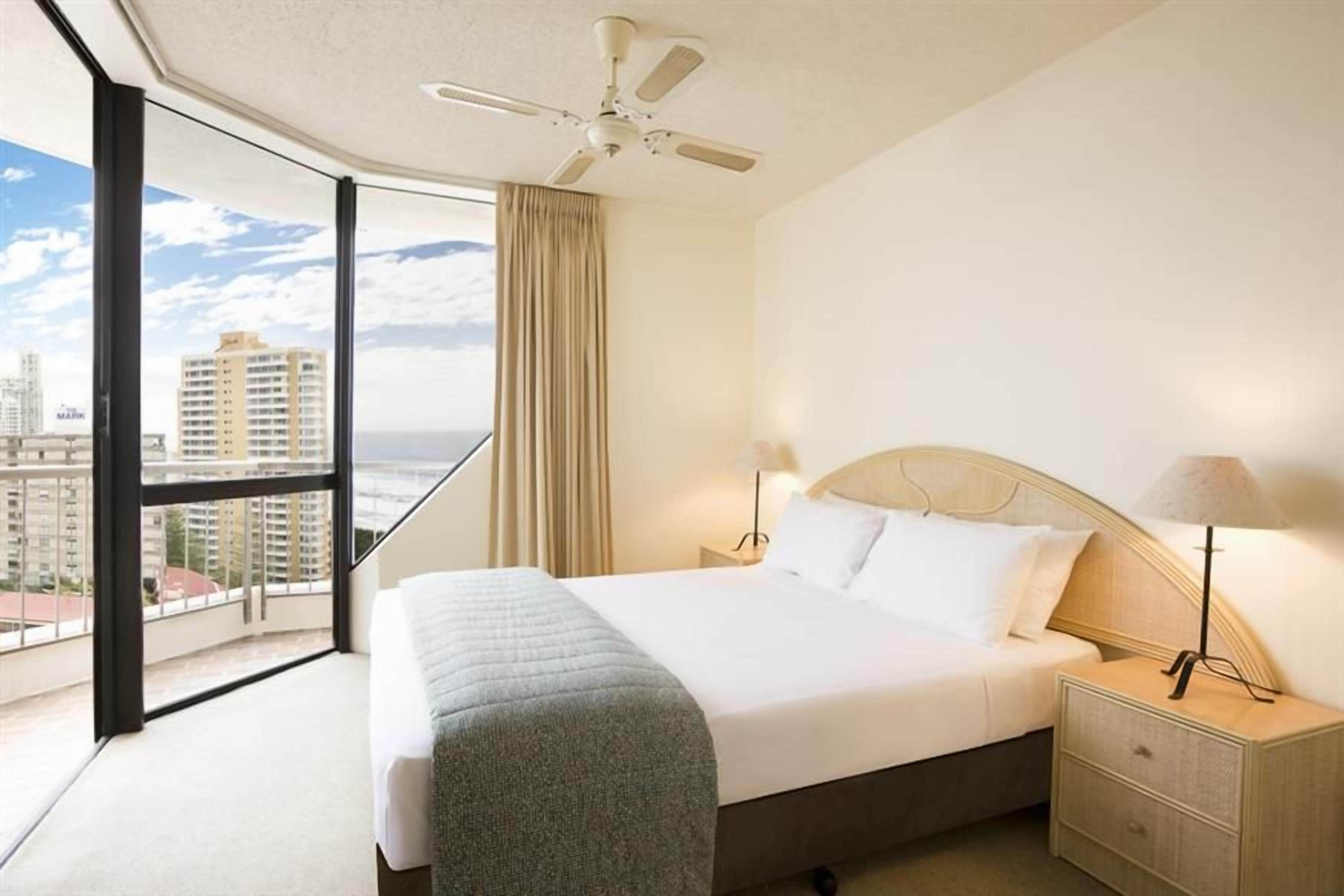 Ein Hotelzimmer mit einem großen Bett, einem Nachttisch, einer Lampe, einem Deckenventilator und Blick auf die Stadt durch ein großes Fenster.
