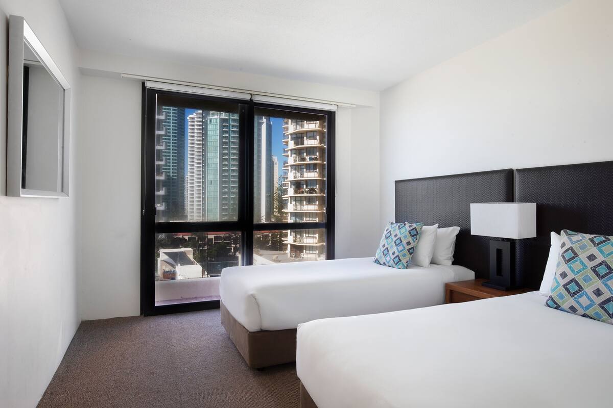 A hotel room with two beds, a large window, and a view of tall buildings.