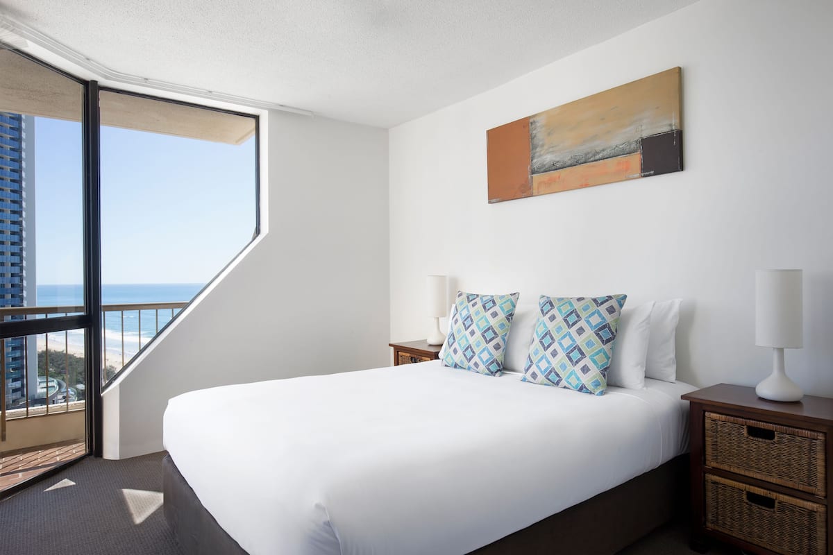A hotel room with a large bed, two nightstands, a wall-mounted painting, and a view of the beach through a large window.