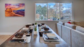 Un coin repas avec une longue table dressée pour le repas, un tableau accroché au mur et une fenêtre donnant sur des arbres et des montagnes.