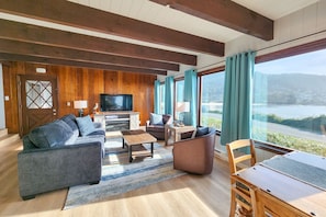 A living room with a large TV, wooden panel walls, a sofa, chairs, and a view of a lake. - Ocean Front 5 BR Home! Yachats, Walk to Everything! Dog Friendly! (Yachats)