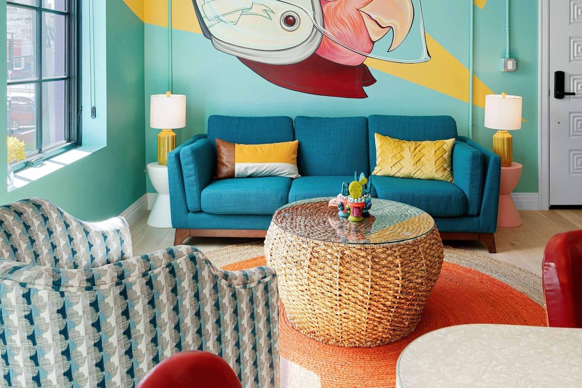 A room with teal walls, a blue sofa, a round wicker coffee table, and a large abstract mural on the wall.