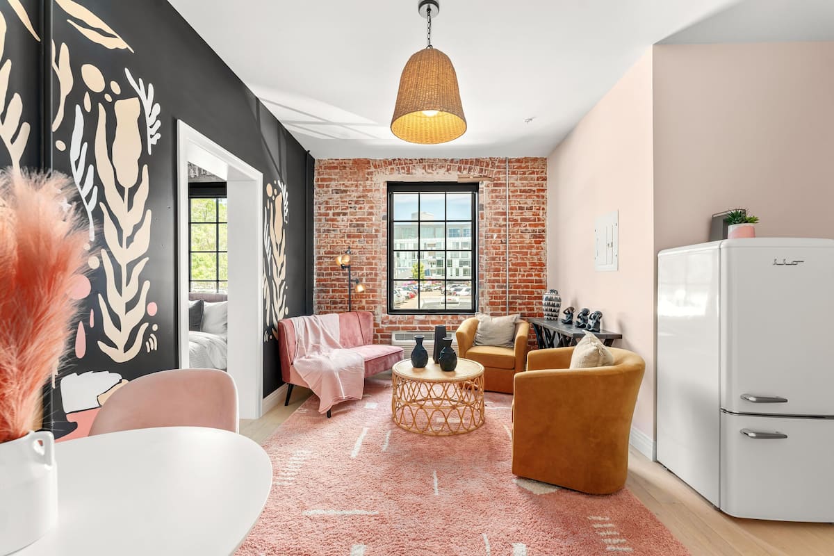 A modern living room with a brick wall, a large window, a white refrigerator, and a pink rug.
