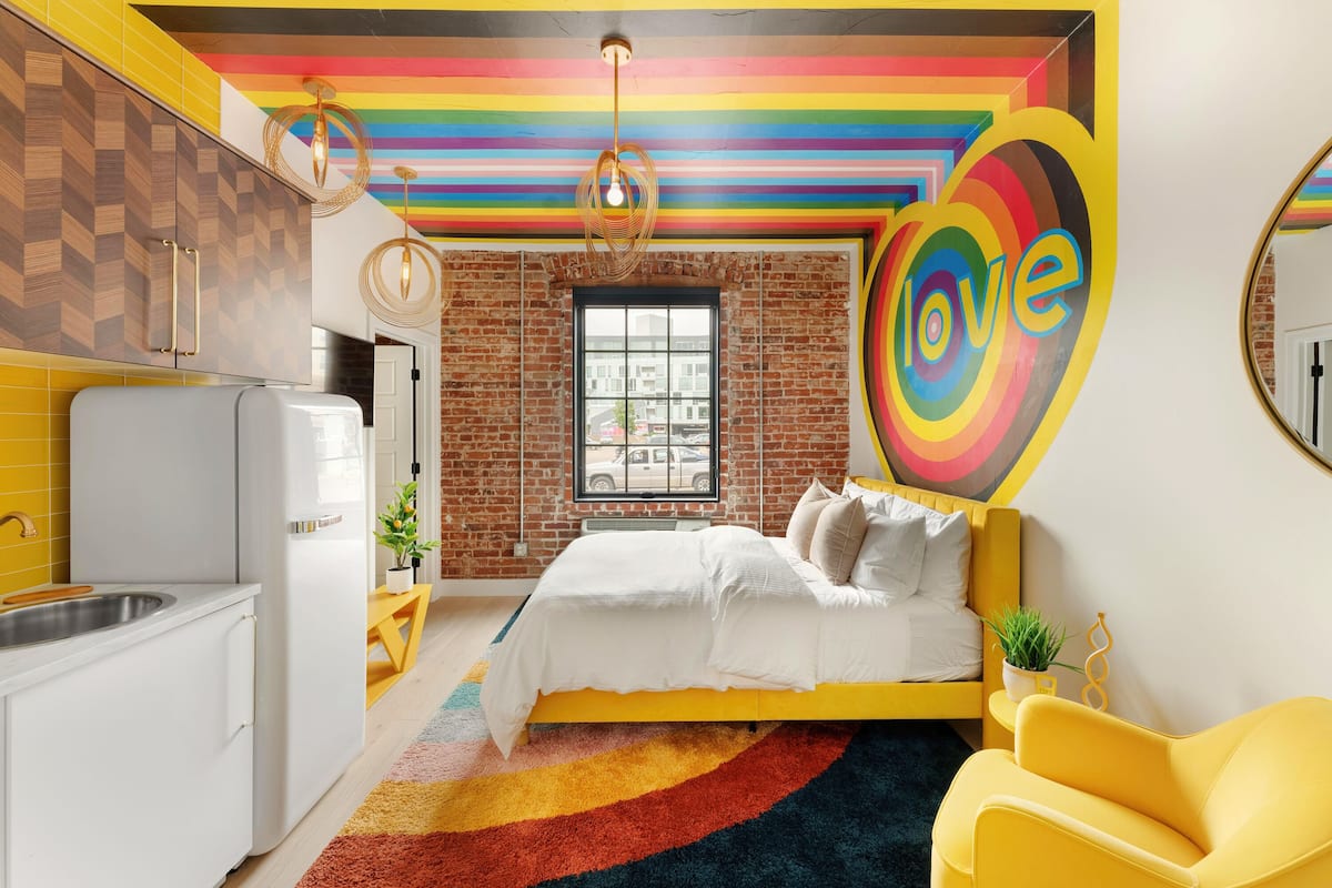 A modern bedroom with a brick wall, a large window, a bed with white bedding, and a colorful rug.