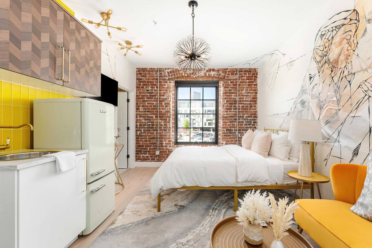 A modern bedroom with a brick wall, a bed, a yellow sofa, and a kitchen area with a refrigerator.