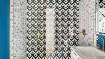 Un baño moderno con baldosas de diseño geométrico, cabina de ducha de vidrio y lavamanos blanco con detalles dorados.