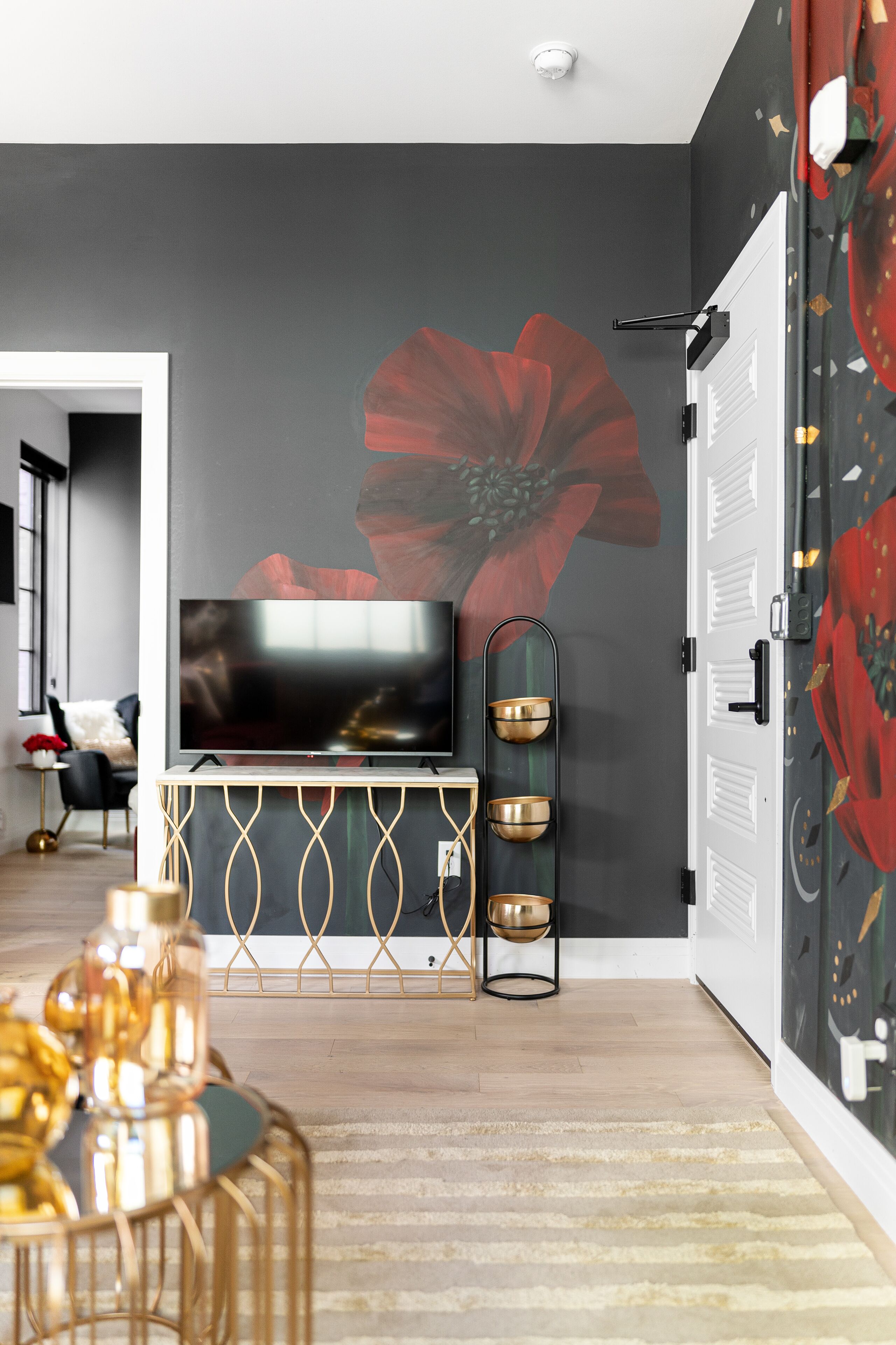 A modern living room with a large flat-screen TV, a decorative wall with a large flower mural, a gold-framed console table, and a striped area rug.
