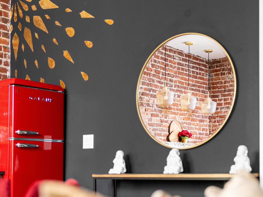 Una habitación con un armario rojo, un espejo redondo que refleja una pared de ladrillo y una mesita con adornos.