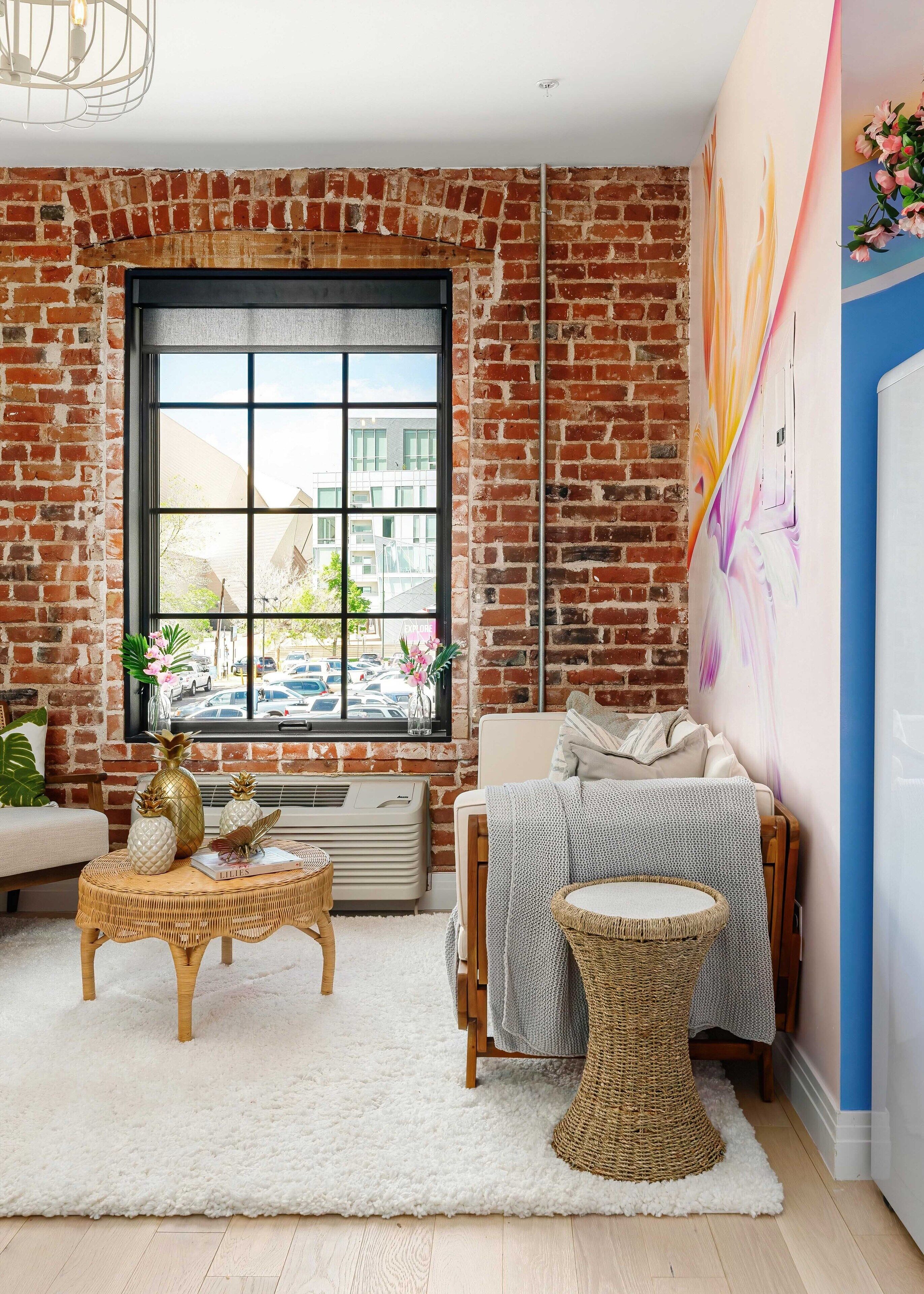 Une pièce chaleureuse avec une grande fenêtre, un mur en briques, un canapé blanc, une table basse en osier, une petite table ronde et une fresque colorée sur le mur.