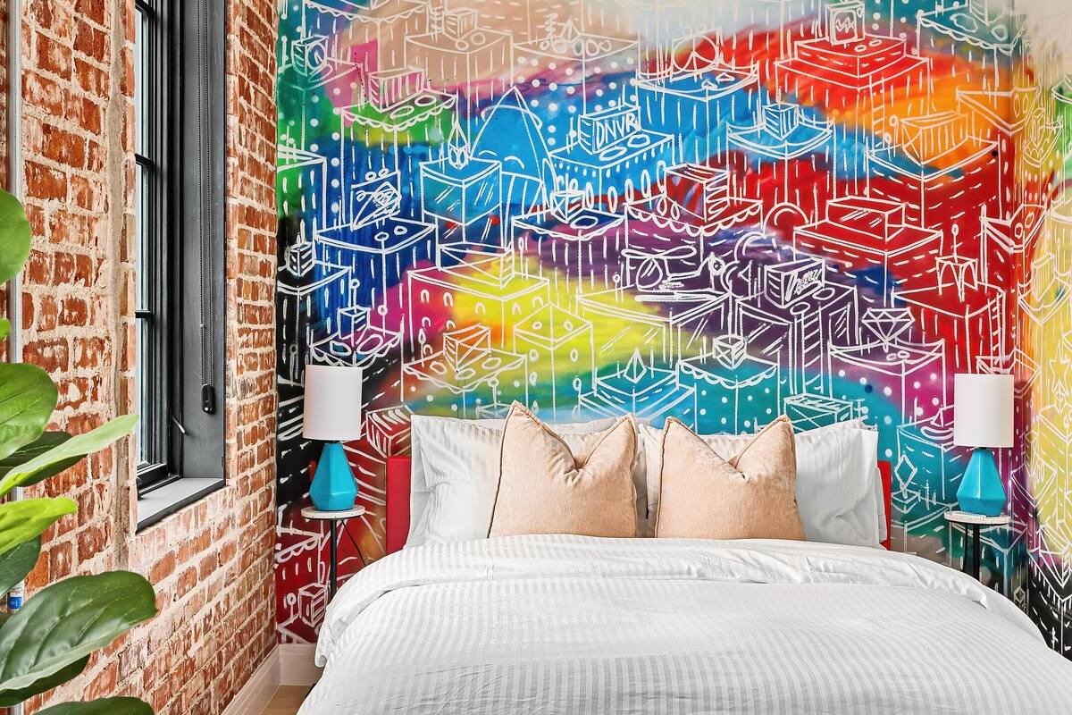 A bedroom with a brick wall, a colorful abstract mural, a bed with white bedding, blue cushions, and two bedside lamps.