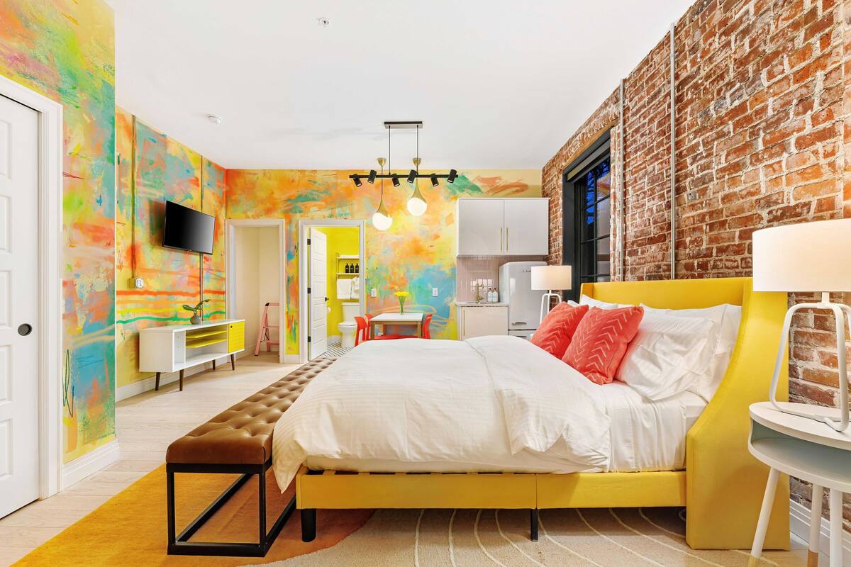 A modern bedroom with a brick wall, a bed with white bedding, a yellow armchair, and a TV mounted on the wall.
