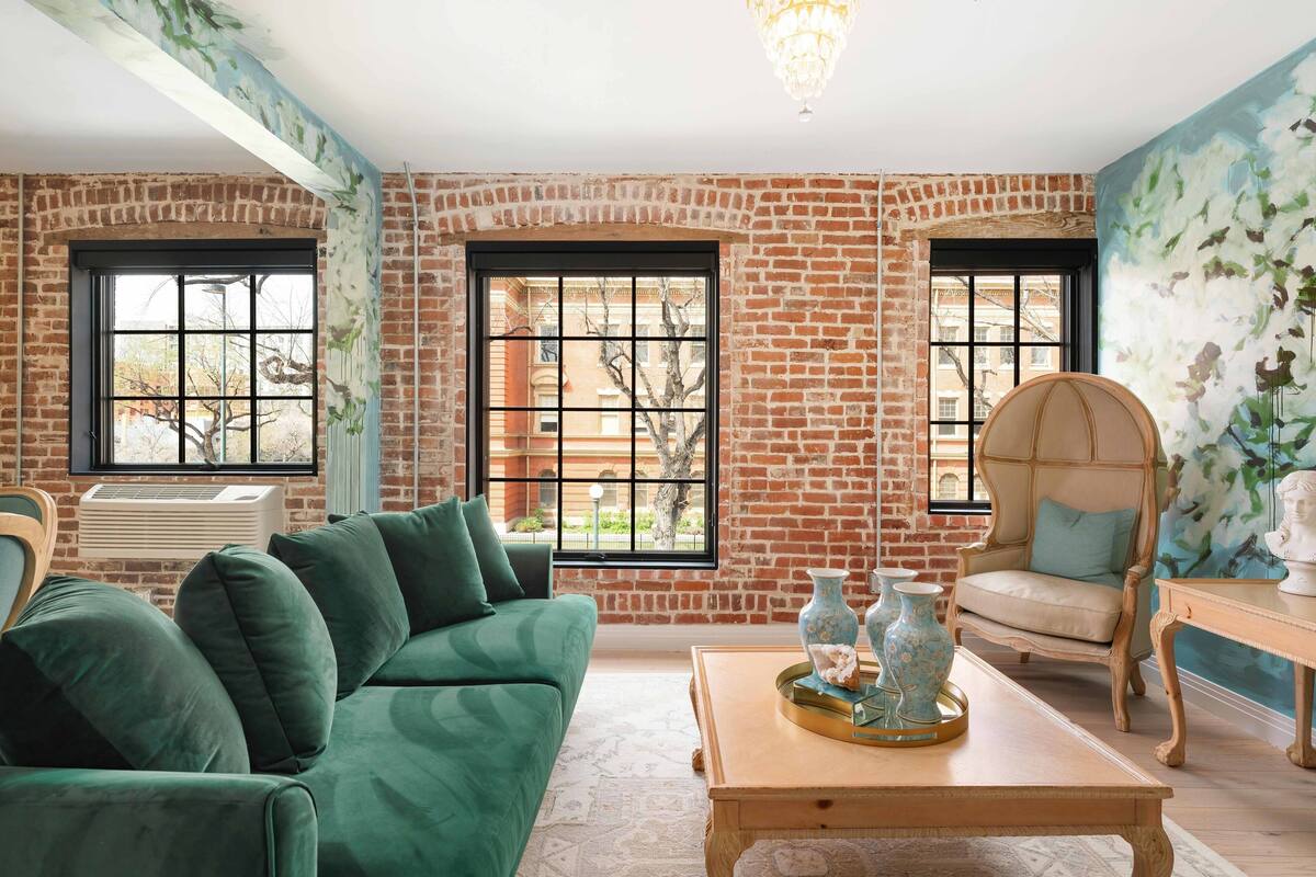 A room with a green sofa, a wooden coffee table with decorative items, a wooden chair, and large windows with a brick wall.