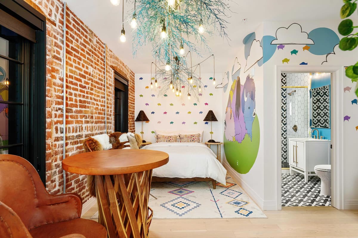 A bedroom with a brick wall, a bed with patterned pillows, a round wooden table, and a hanging light fixture.