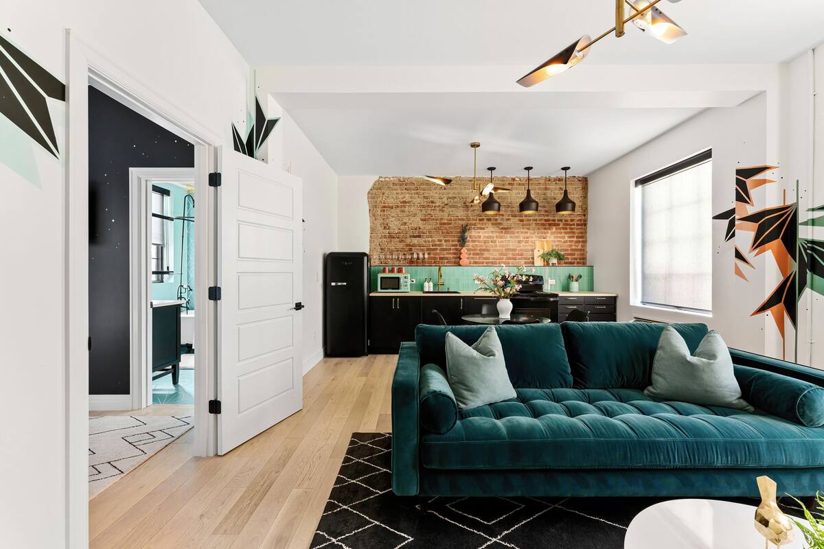 A modern living room with a teal sofa, a black and white rug, and a kitchen with a brick wall and pendant lights.