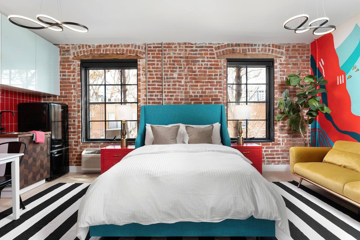 A modern bedroom with a brick wall, a teal headboard, a large bed, a yellow sofa, and a red nightstand.