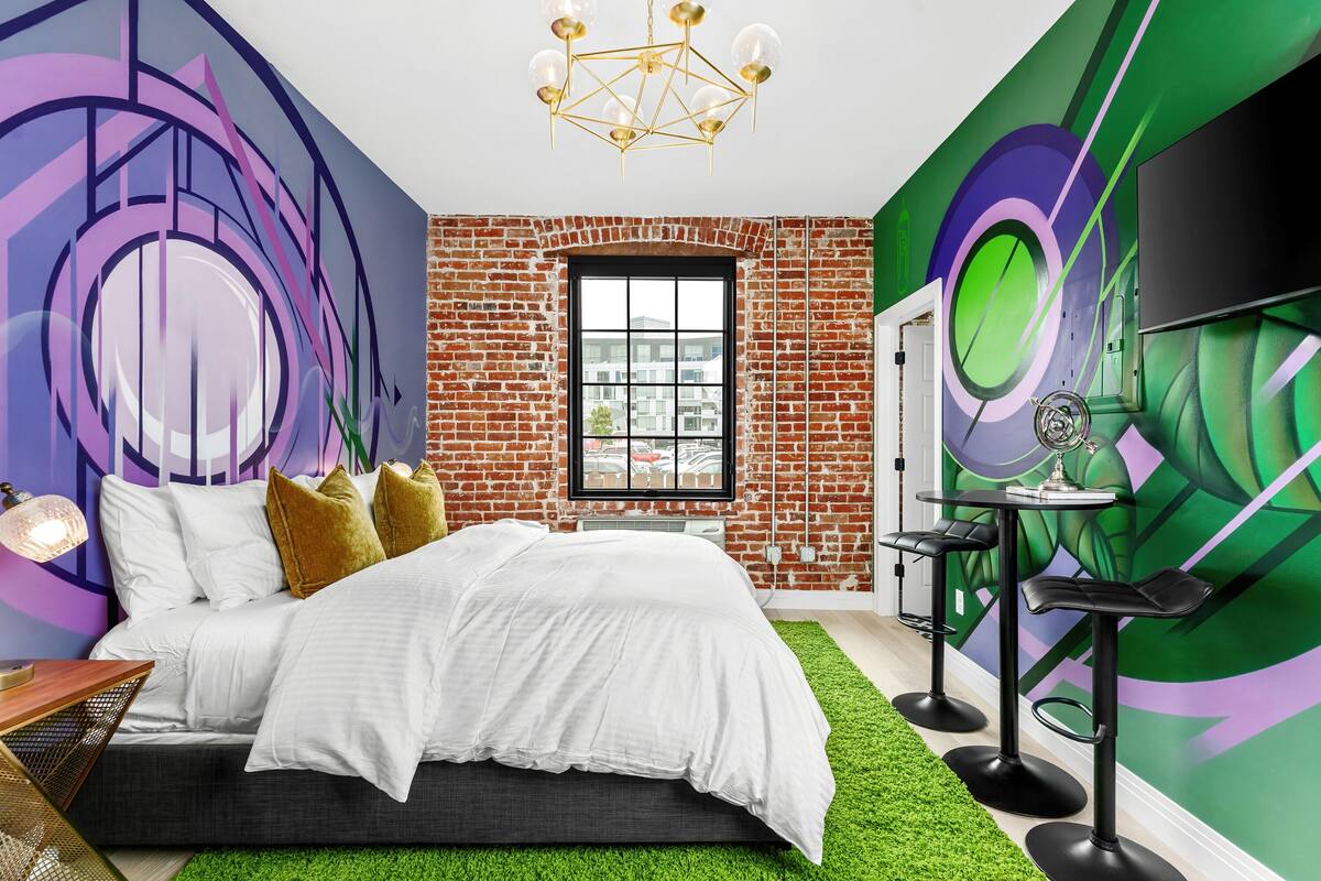 A modern bedroom with a brick wall, a large window, a bed with white bedding, a green rug, and a TV mounted on the wall.