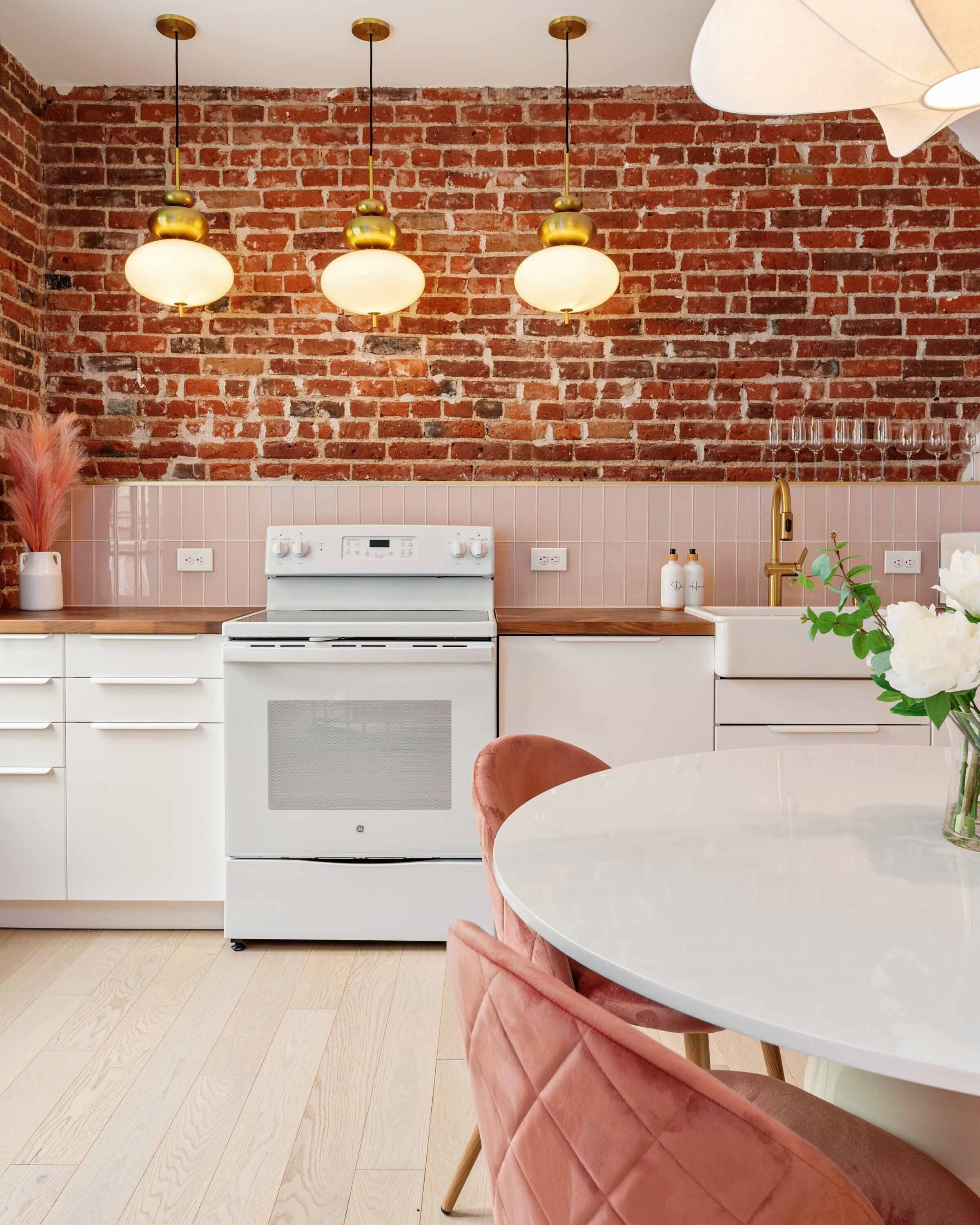 Une cuisine avec un mur en briques, des meubles blancs, une cuisinière blanche et des chaises roses.